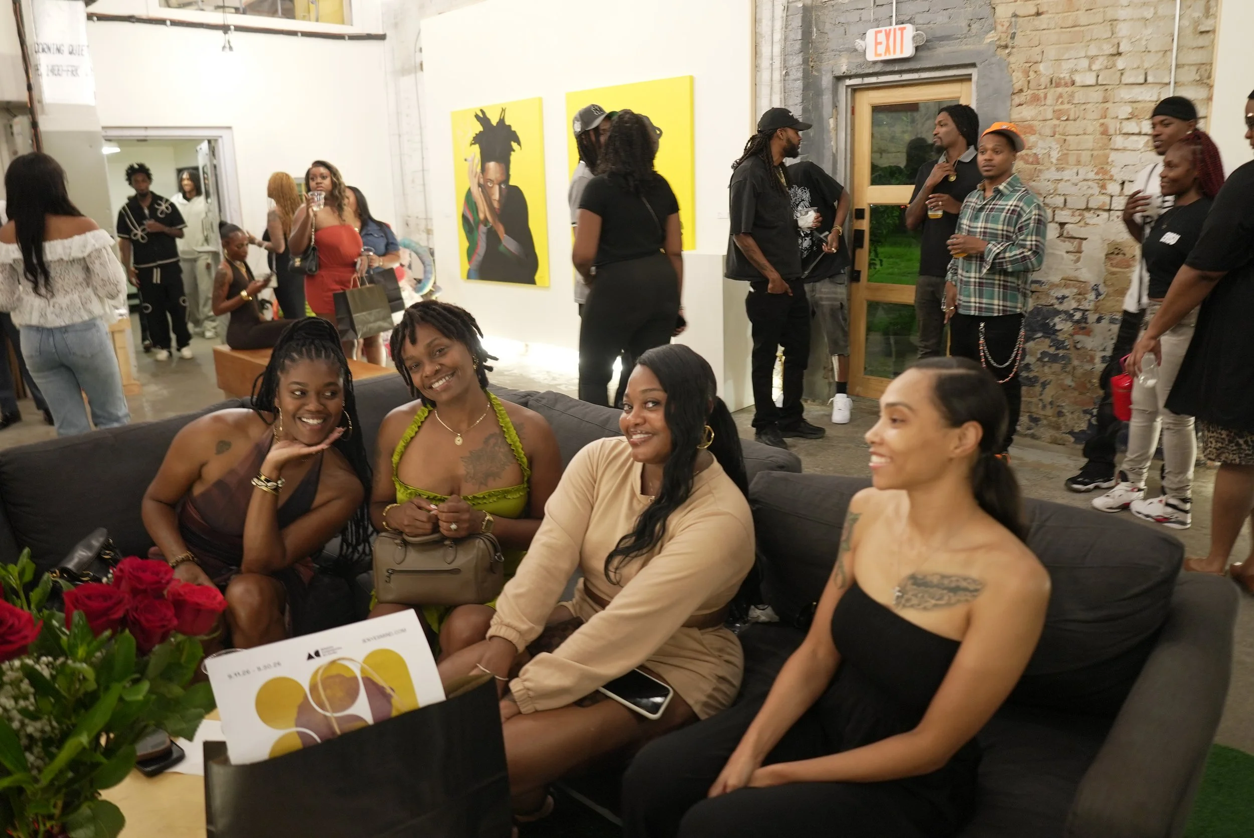 People gathering at an indoor art gallery or event space, socializing and sitting on couches, with artwork on the walls and an exposed brick wall on one side.