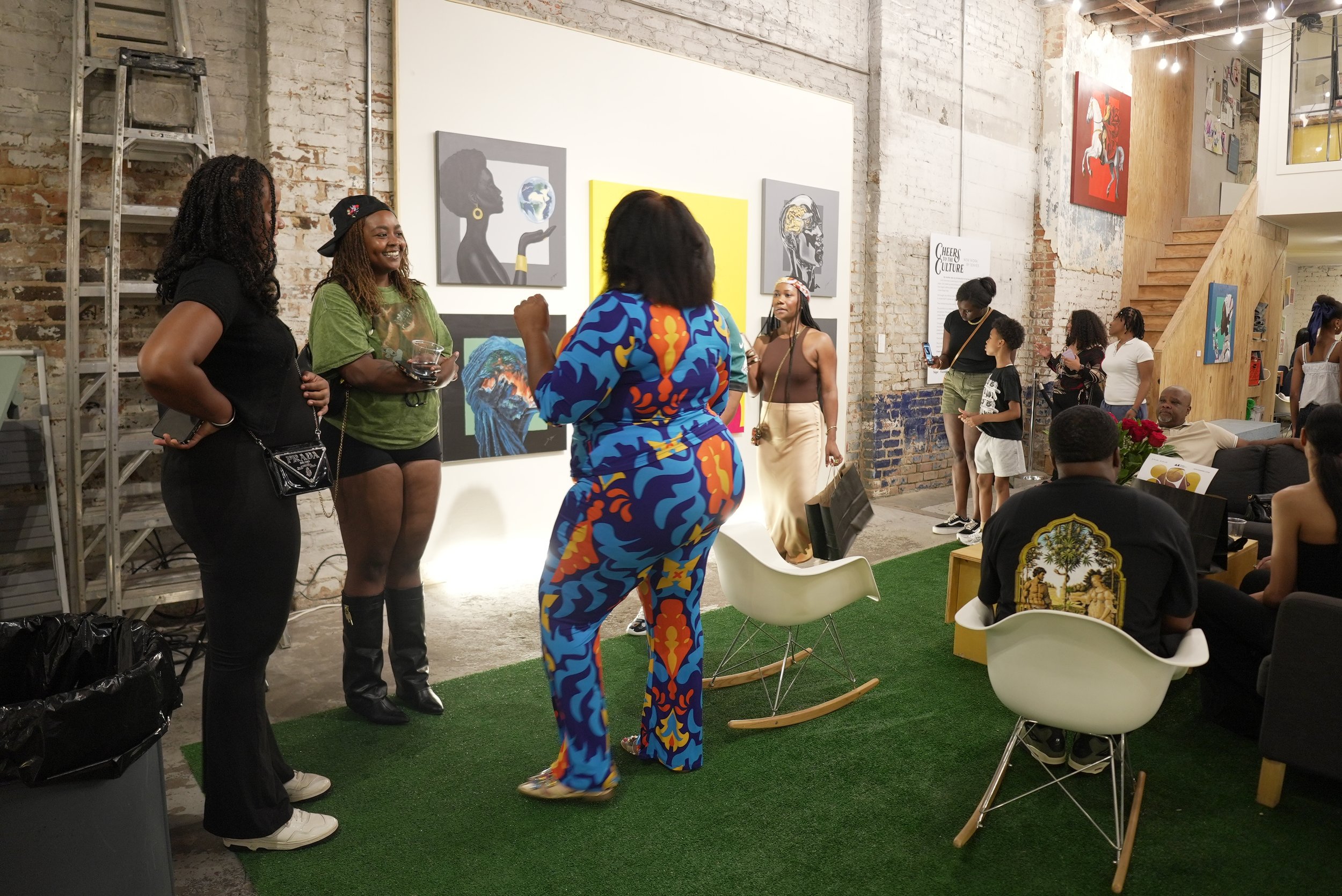 People socializing inside an art gallery with colorful paintings on brick walls, some individuals are taking photos and engaging in conversation.