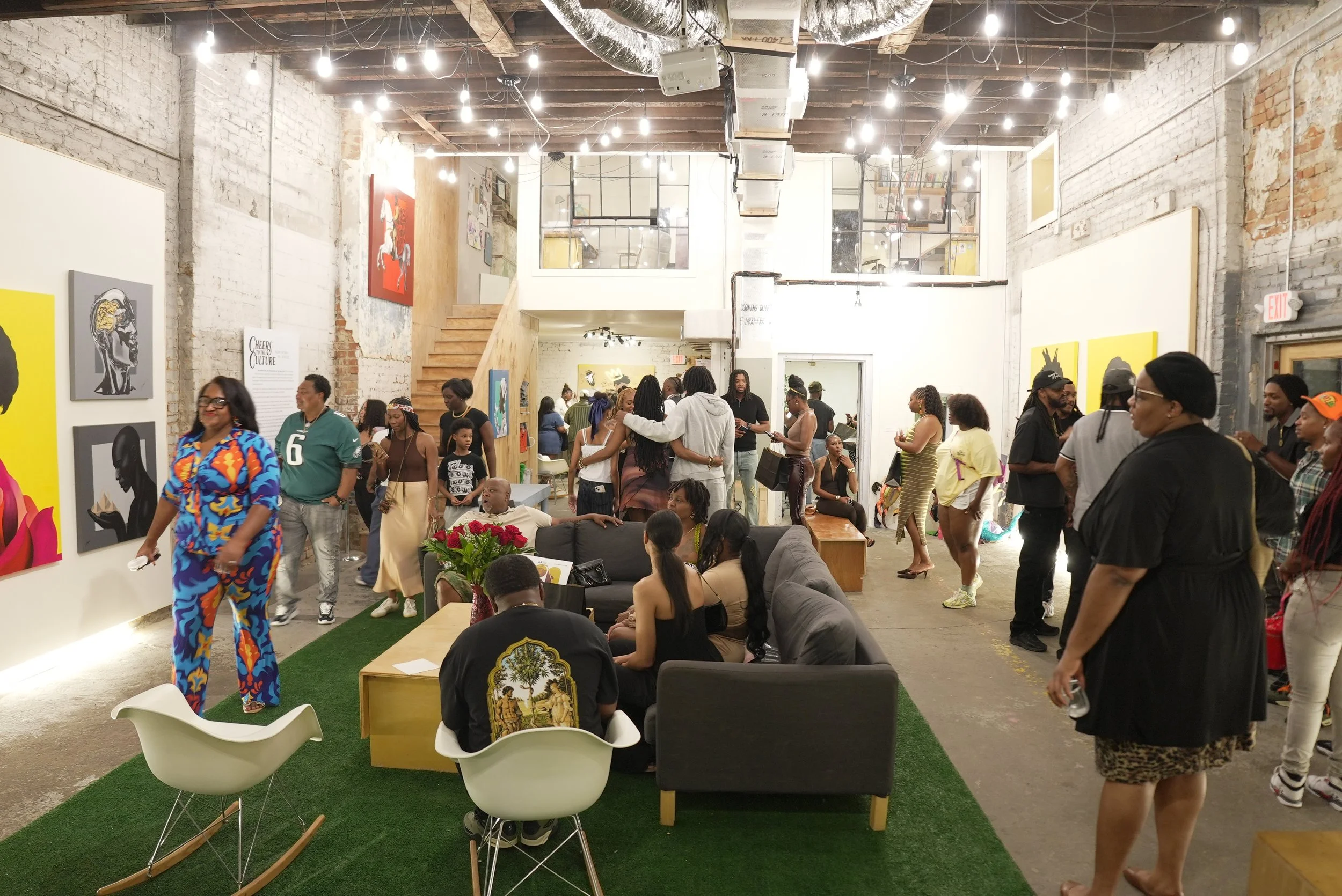 People socializing in an art gallery with abstract paintings on white brick walls and exposed ceiling beams, some sitting on couches and others standing and walking around.