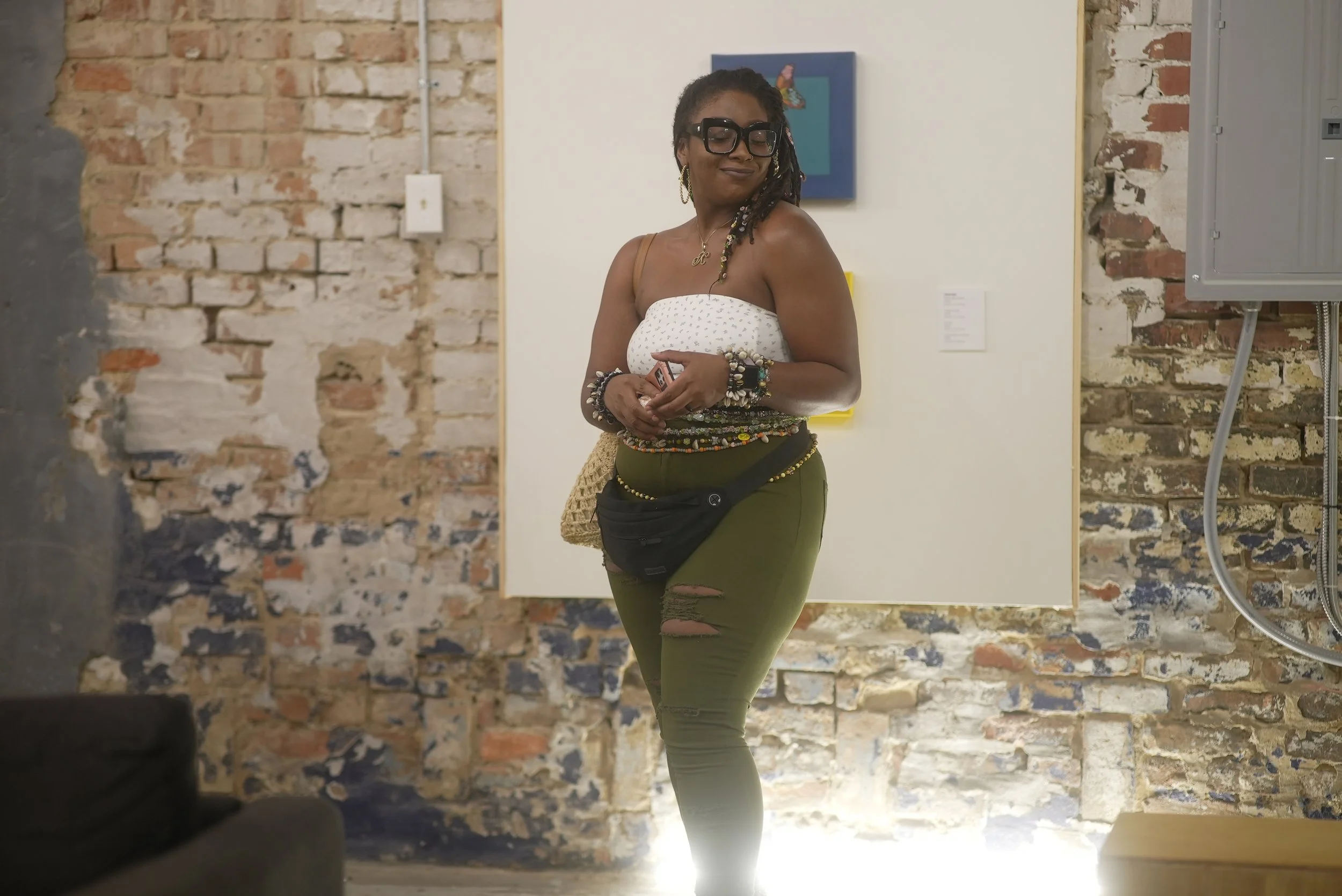 A woman with dreadlocks wearing glasses, a white strapless top, and green ripped jeans, holding a phone, standing against exposed brick and white walls in an art gallery or studio.