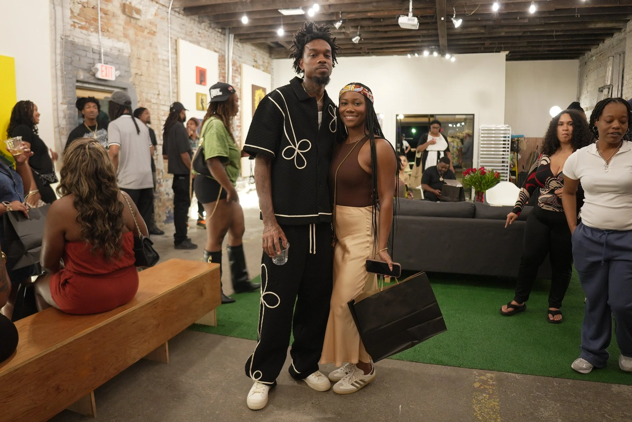 A group of people gathered in an art gallery with exposed brick walls and hanging artworks. Two people are standing in the center, smiling at the camera, holding shopping bags and drinks, with other attendees in the background.