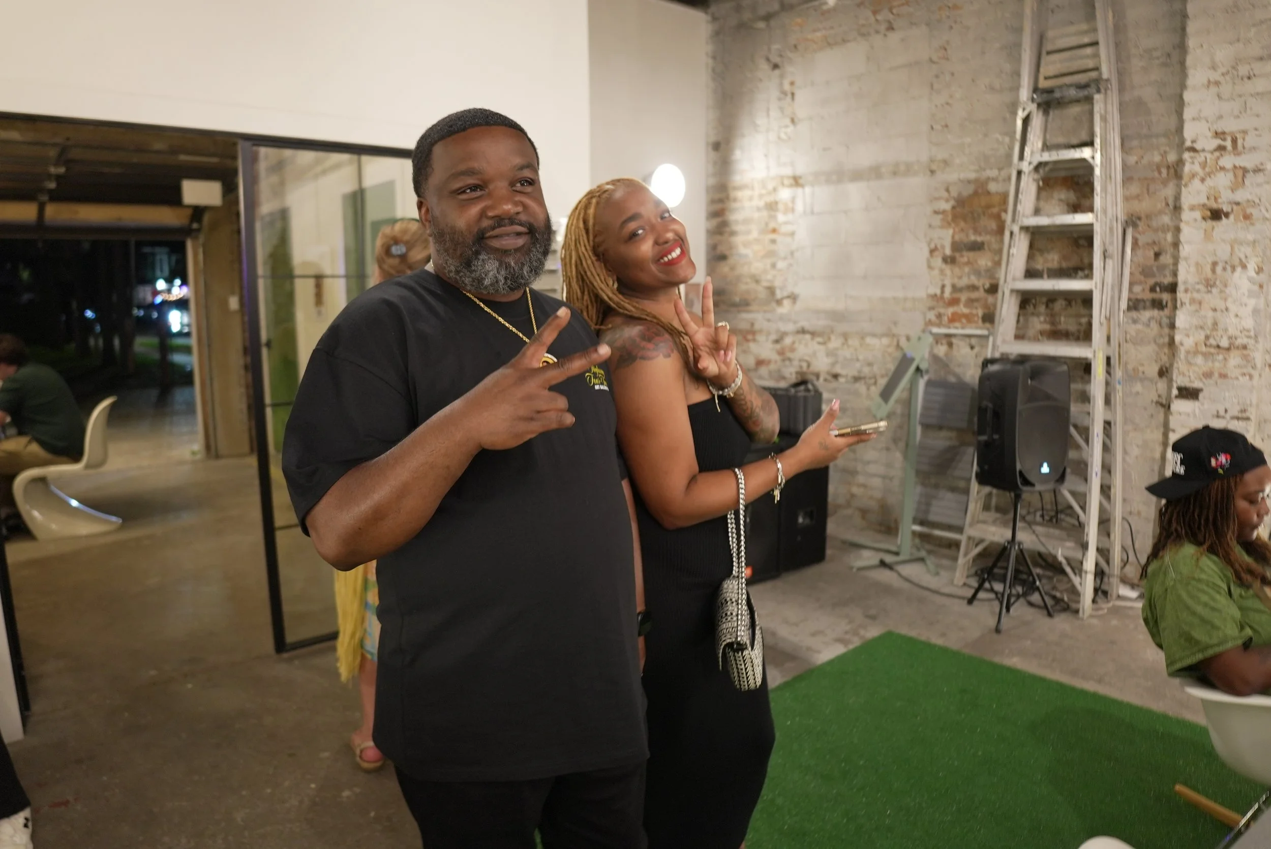 Two people posing indoors, one man and one woman, both making peace signs and smiling, standing near a ladder and a speaker.