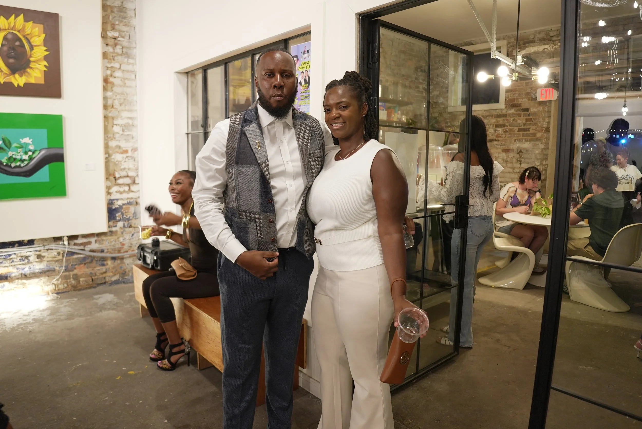 A man and woman standing together inside an art gallery, smiling at the camera. The gallery has exposed brick walls and various artworks hanging.