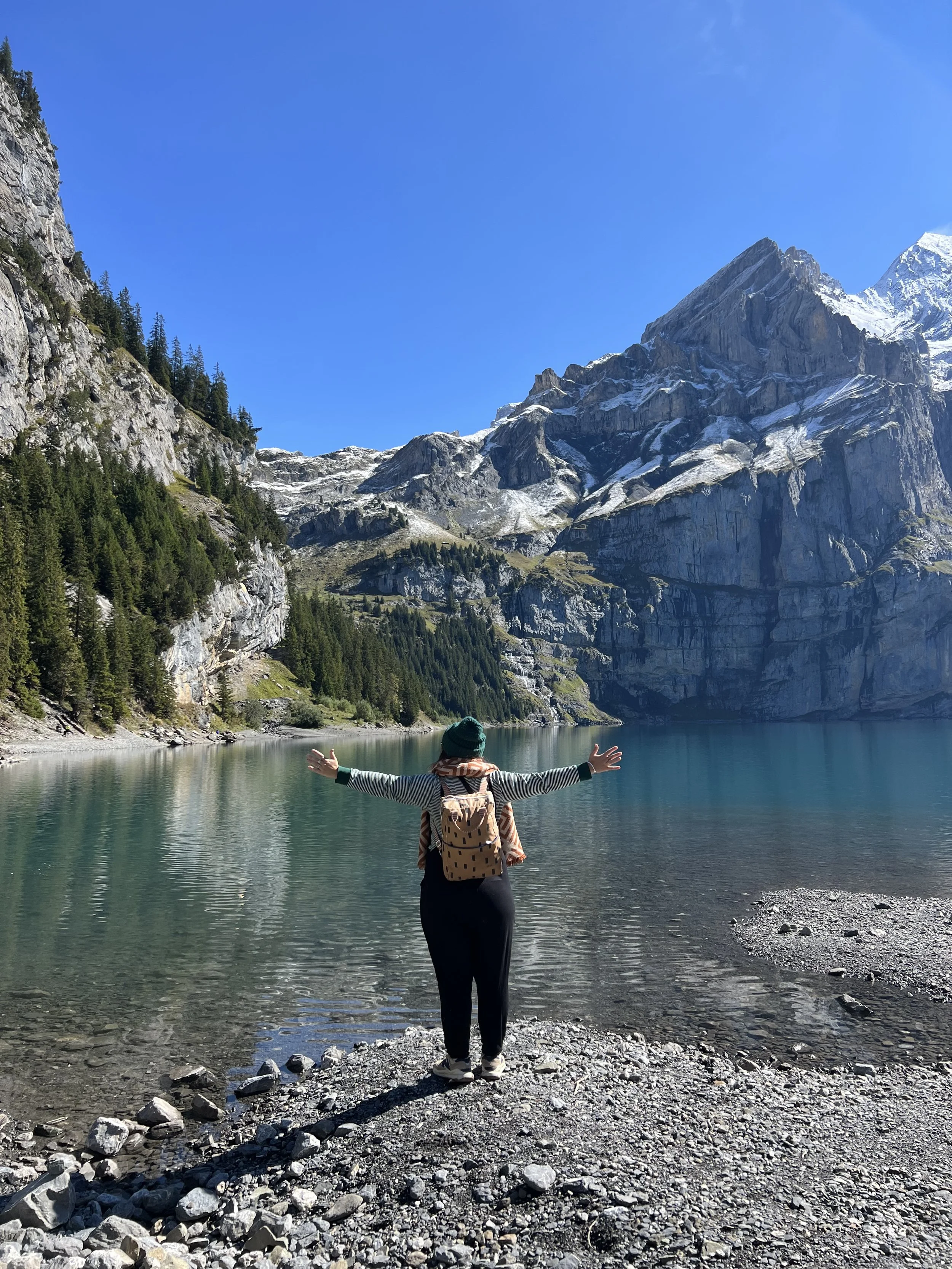 Person standing at the edge of a lake with arms outstretched, surrounded by tall mountains with snow on the peaks and a clear blue sky.