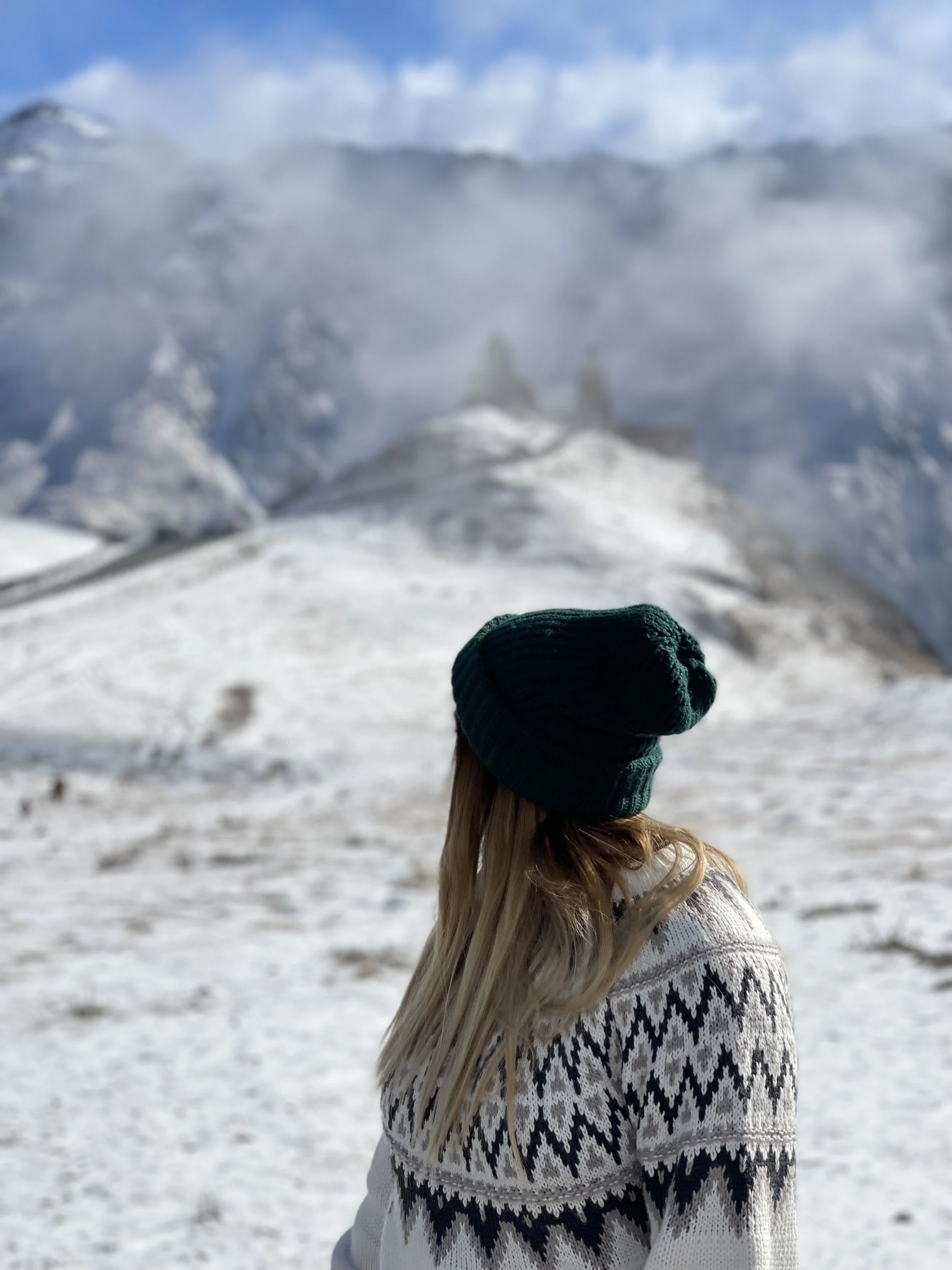 A woman with long blonde hair wearing a green knit hat and a patterned sweater stands in a snowy landscape, with snow-covered mountains and partly cloudy sky in the background.