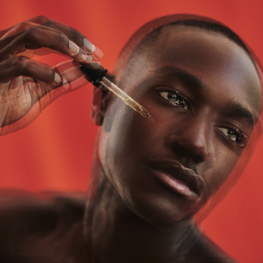 A person with dark skin and short hair holds a dropper near their face, with a neutral expression and a red background.