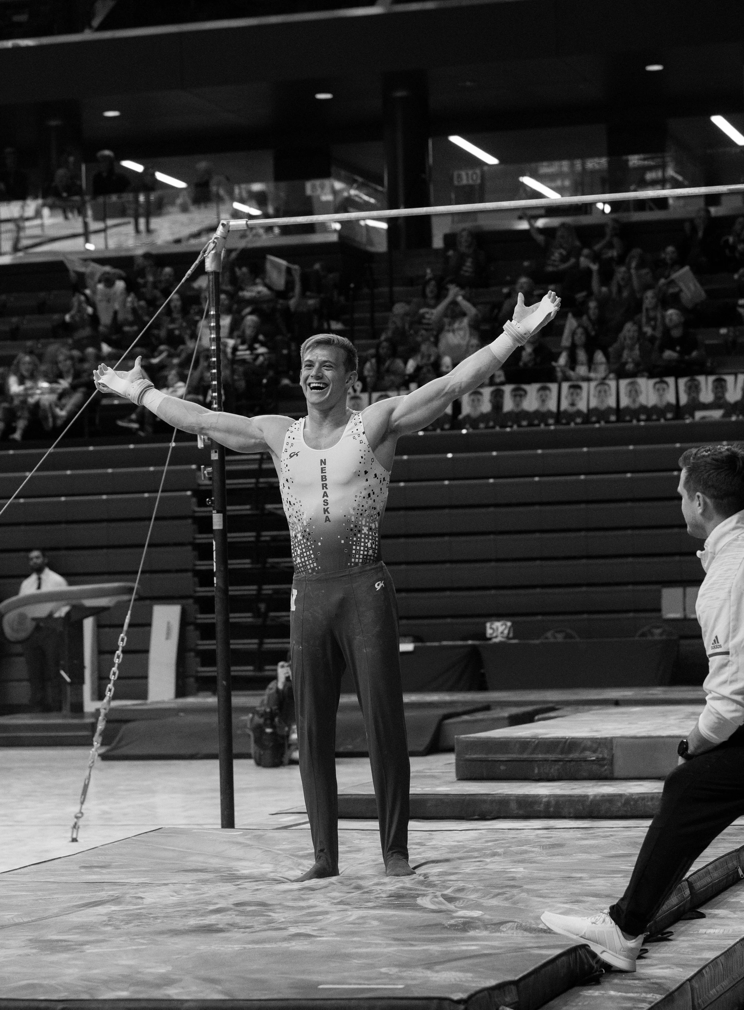 A male gymnast stands on the mat with arms outstretched, smiling, during a gymnastics competition. He is wearing a sleeveless leotard with 'NEBRASKA' written down the front, with an audience and judges visible in the background.