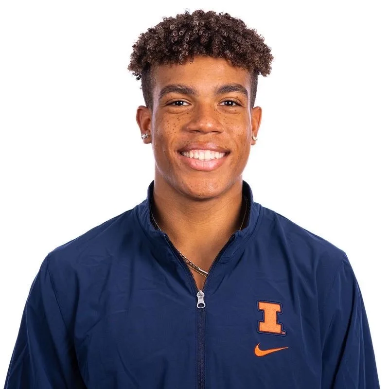 Young man with curly hair smiling, wearing a navy blue sports jacket with an orange 'I' logo and Nike swoosh.