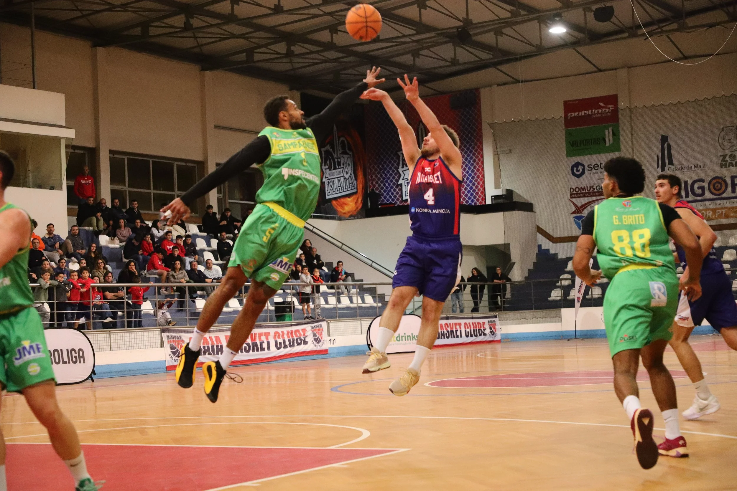 A basketball game in progress with players jumping for the ball, spectators watching from the stands.