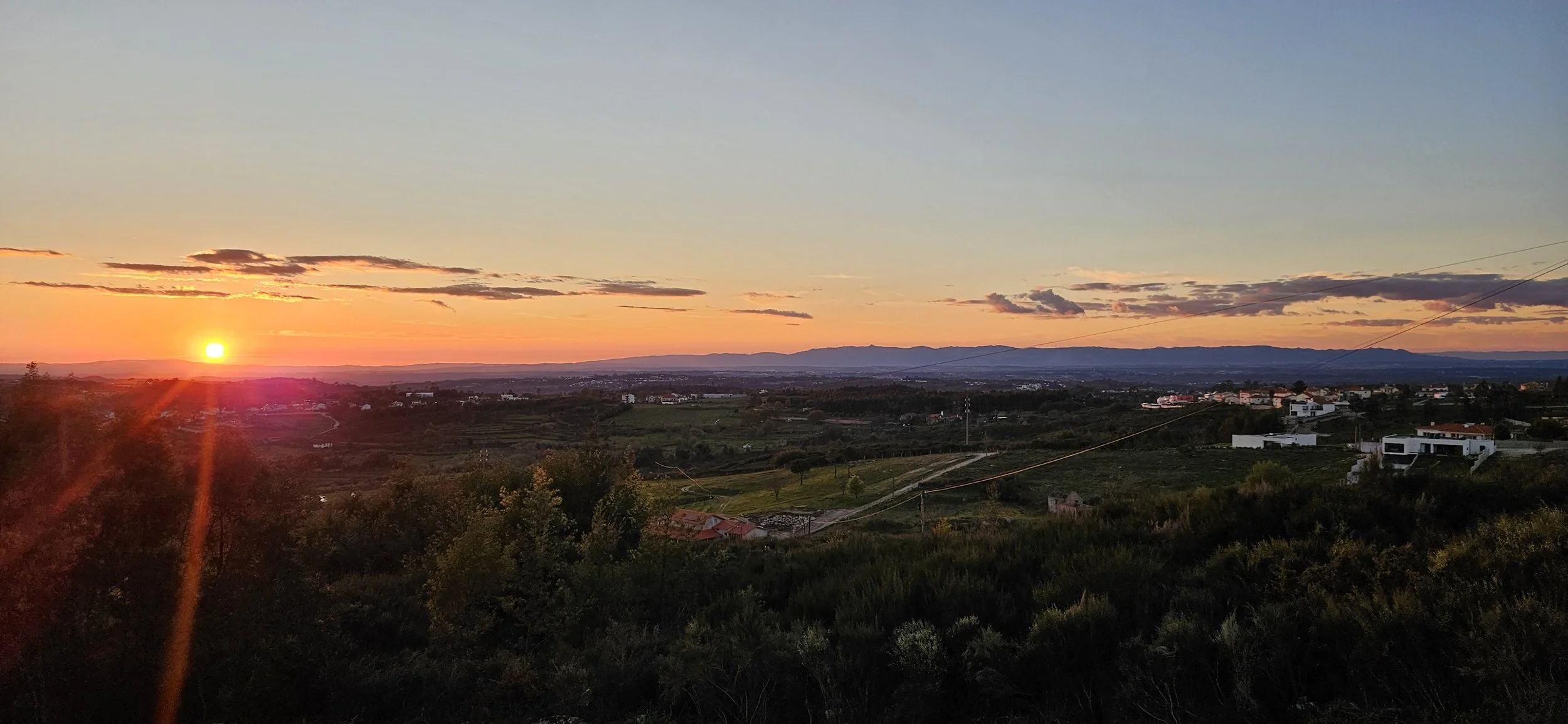 Sunset over a rural landscape with fields, trees, and distant houses under a partly cloudy sky.