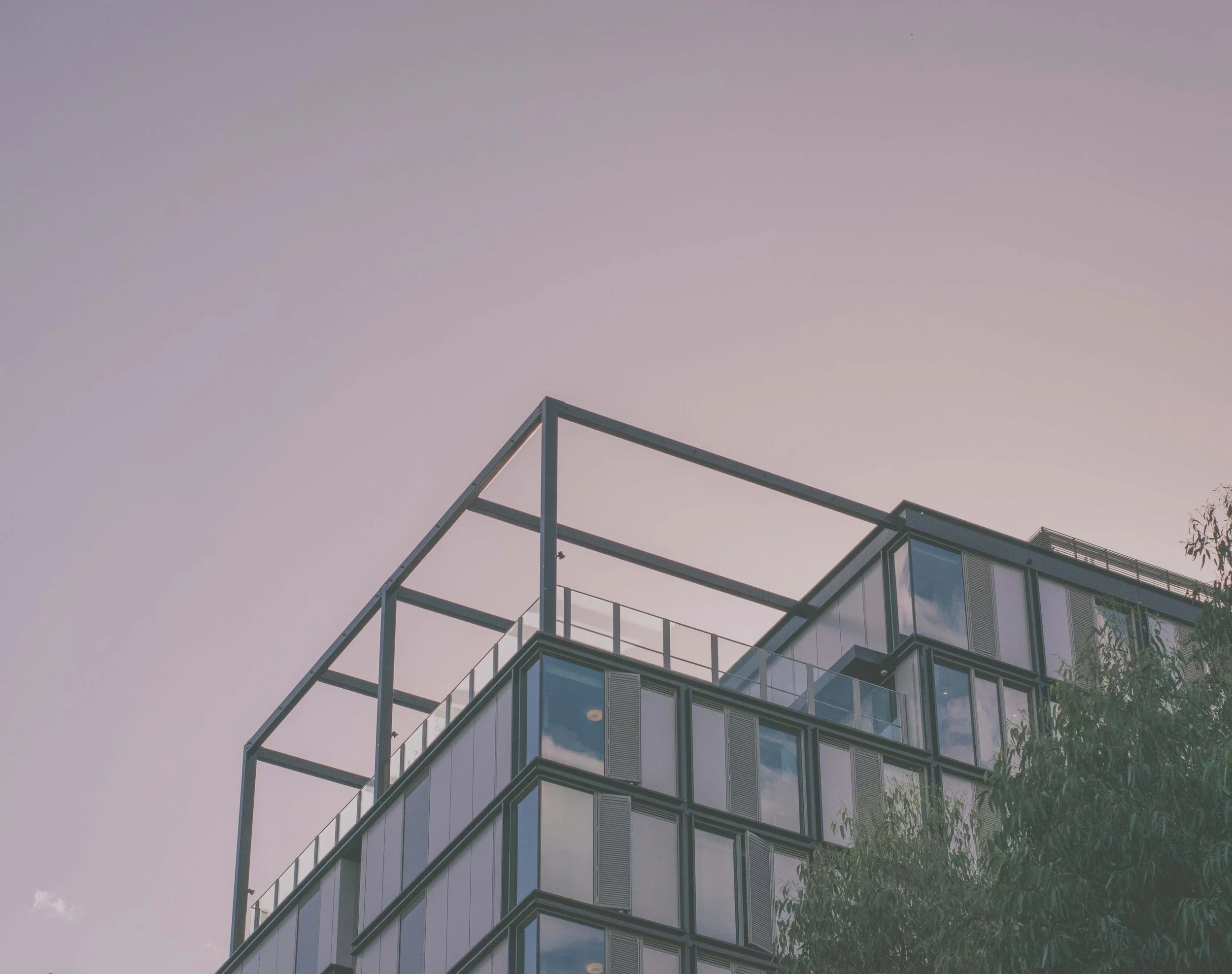 Modern multi-story building with glass windows and metal framing, sky and clouds reflected in the windows, and a tree in the foreground.