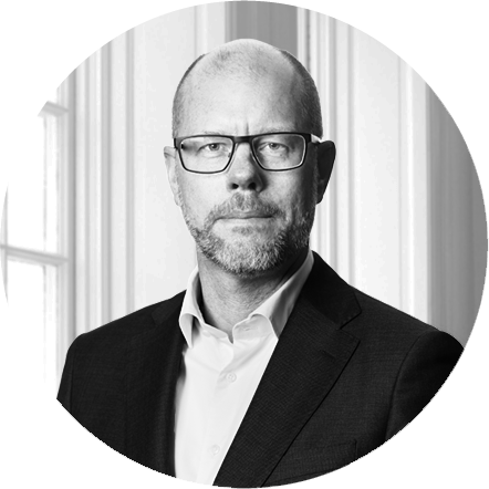 Black and white portrait of a middle-aged man with glasses, wearing a suit and white shirt, standing indoors in front of large windows.