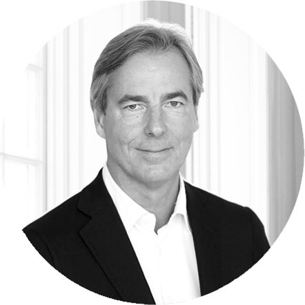 A professional black-and-white portrait of a middle-aged man with gray hair, wearing a black suit jacket and white shirt, standing in front of vertical blinds.