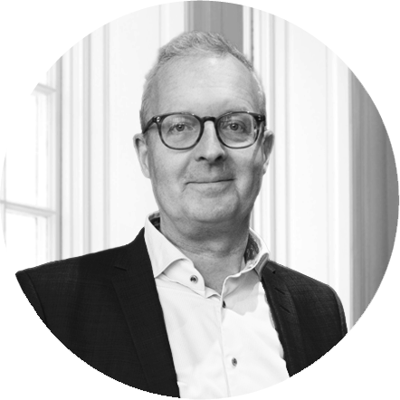 Black and white portrait of a man wearing glasses, a white dress shirt, and a dark blazer, standing indoors in front of a window with vertical blinds.