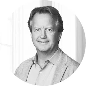 Black and white portrait of a middle-aged man with light hair, wearing a blazer over a collared shirt, smiling gently, with a blurred background.