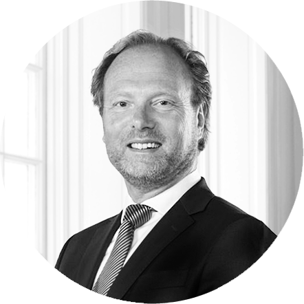 Black and white portrait of a man in a suit and tie, smiling, standing in front of large windows.