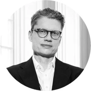 Black-and-white portrait of a woman with short hair, glasses, wearing a suit jacket and collared shirt, standing in front of a window.