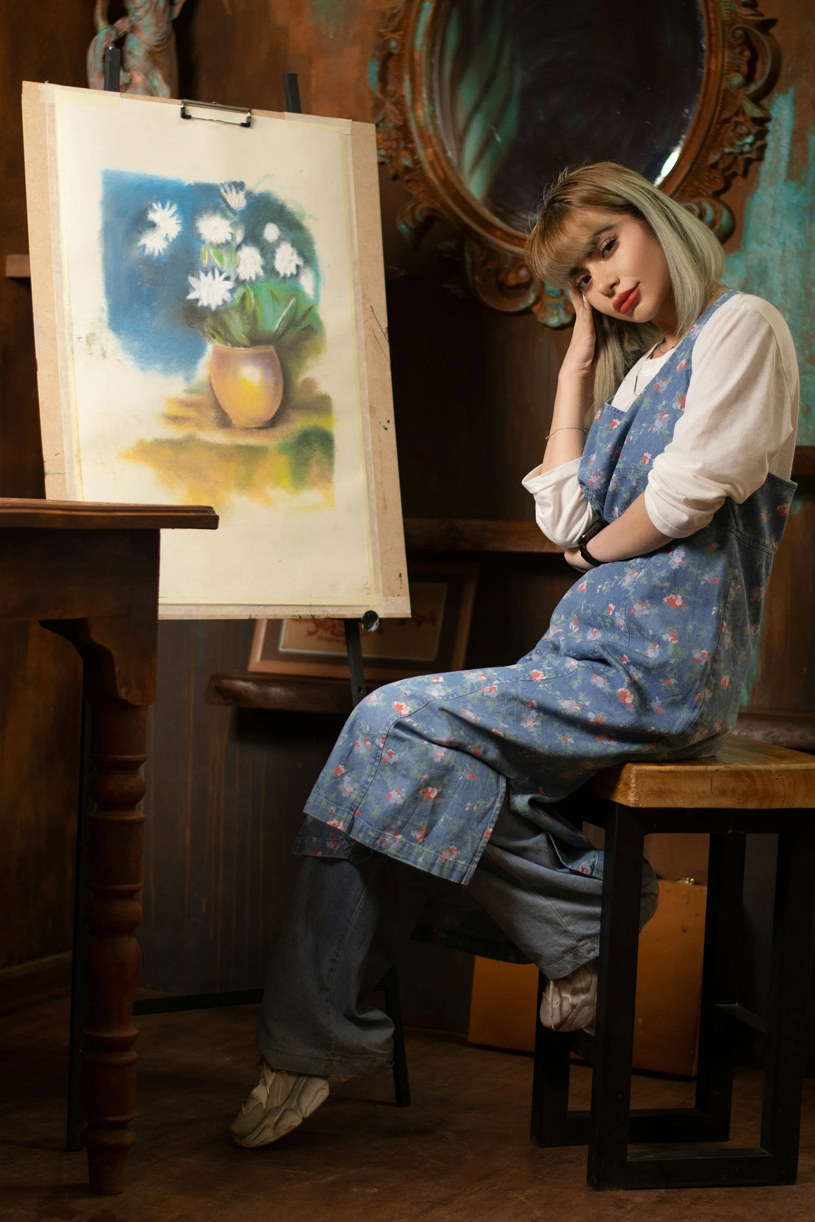 A young woman with light-colored hair sitting on a wooden bench in an art studio, wearing a denim apron over a white shirt, with a watercolor painting of daisies on an easel beside her.