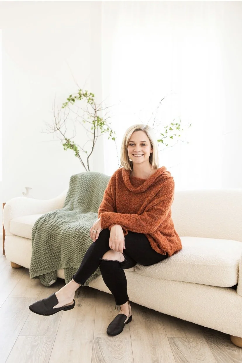 A smiling woman with blonde hair sitting on a cream-colored sofa in a bright room. She is wearing a rust-colored sweater, ripped black jeans, and black slip-on shoes. There is a green blanket draped over the arm of the sofa and a small tree with green and bare branches behind her.