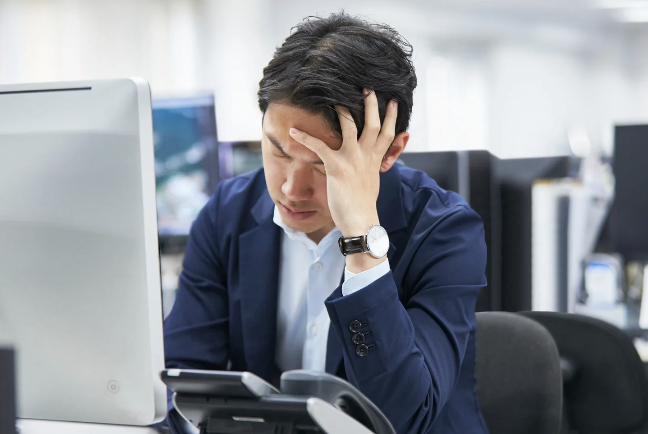 Man in business attire sitting at a desk with a computer and phone, holding his head in frustration or stress.