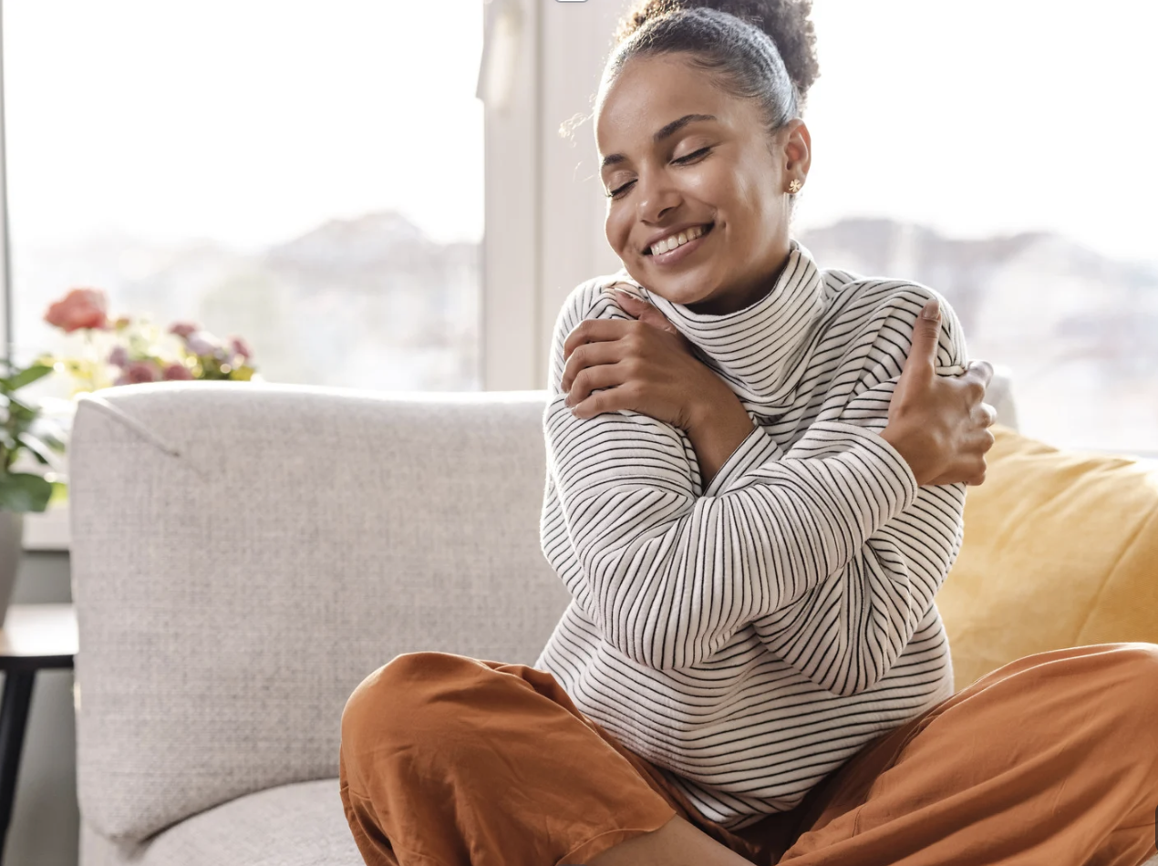 A woman sitting on a couch hugging herself with a smile, wearing a striped turtleneck and brown pants, in a cozy, well-lit room with large windows and a potted plant in the background.