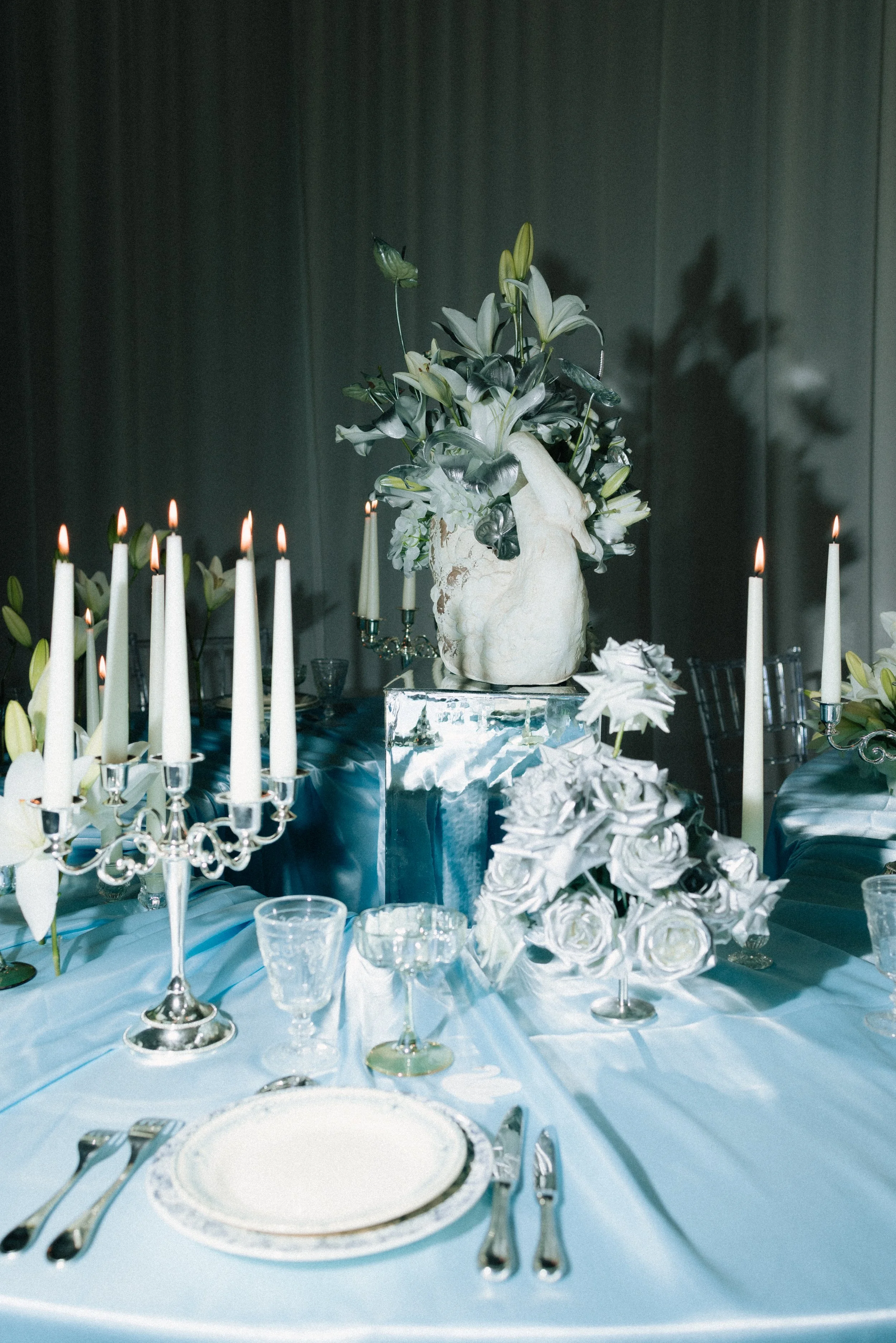 Table de banquet décorée avec des bougies allumées, un vase avec des fleurs blanches et vertes, et diverses assiettes et couverts, dans une ambiance élégante et sophistiquée.