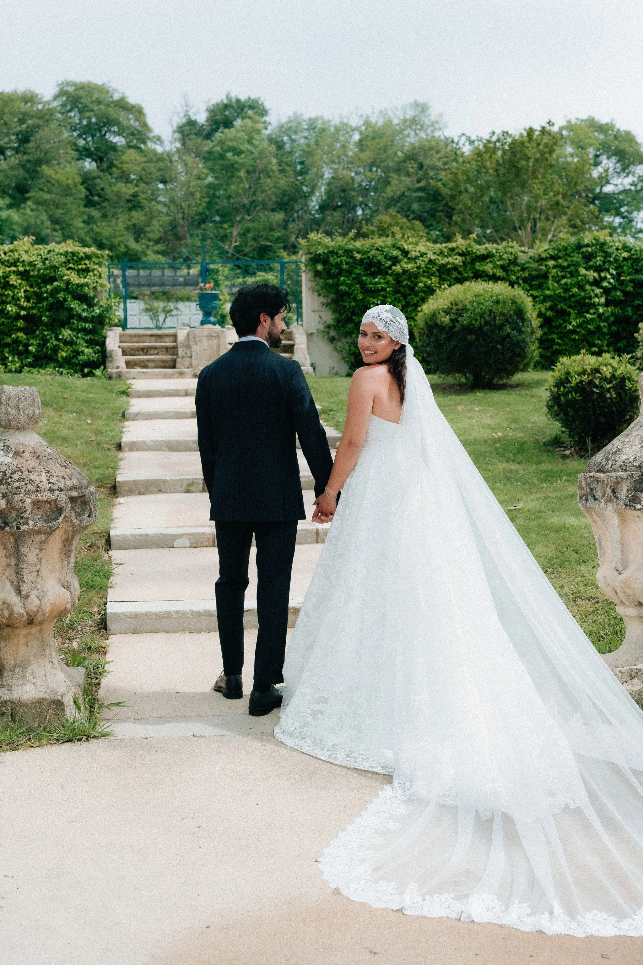 Un couple de mariés marche main dans la main dans un jardin verdoyant, avec un escalier en pierre derrière eux, entouré d'arbres et de buissons, lors d'une journée ensoleillée.