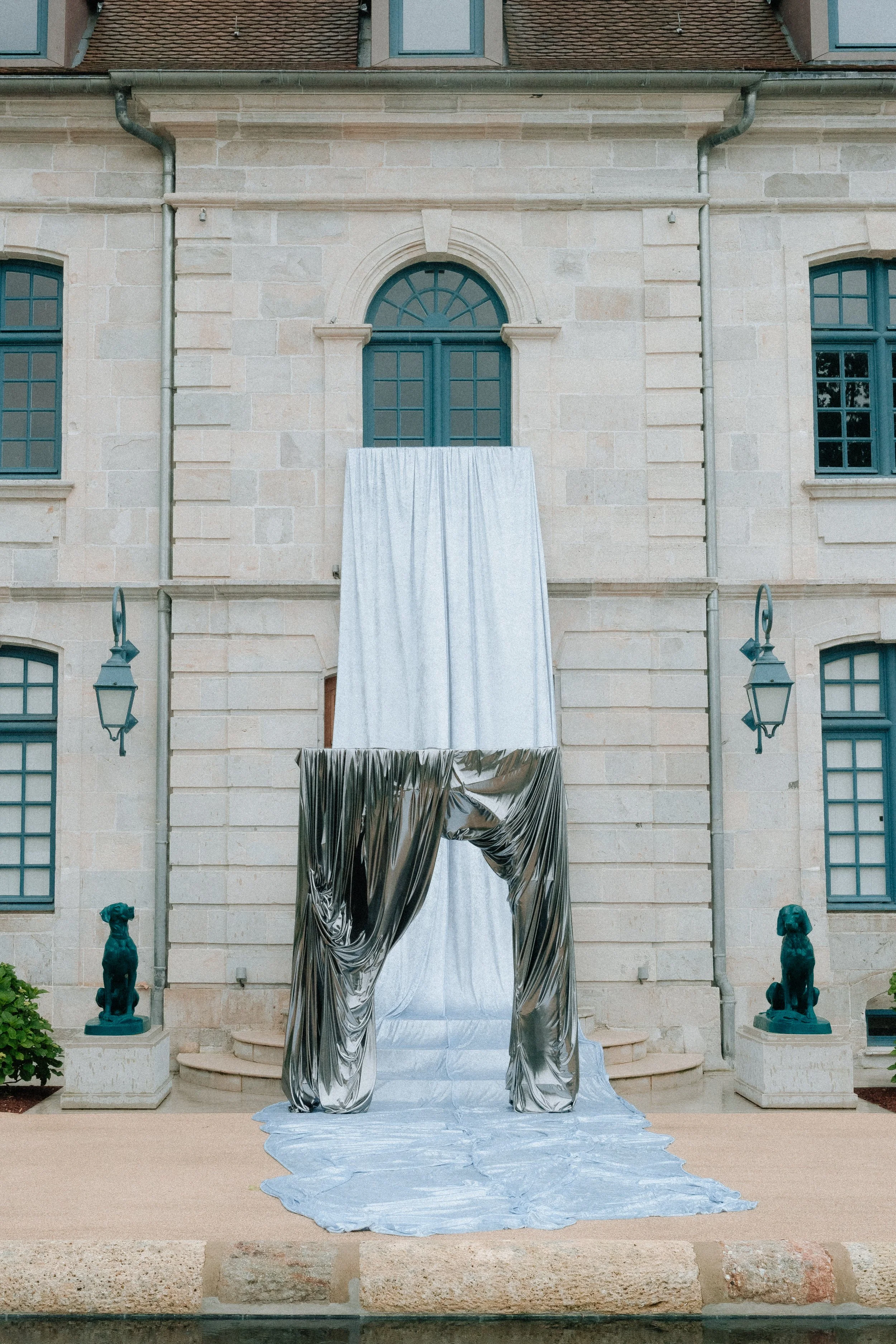 Installation artistique avec un tissu blanc et des tissus métalliques argentés devant une façade en pierre d'une maison, avec deux statues de chiens en bronze à chaque côté.