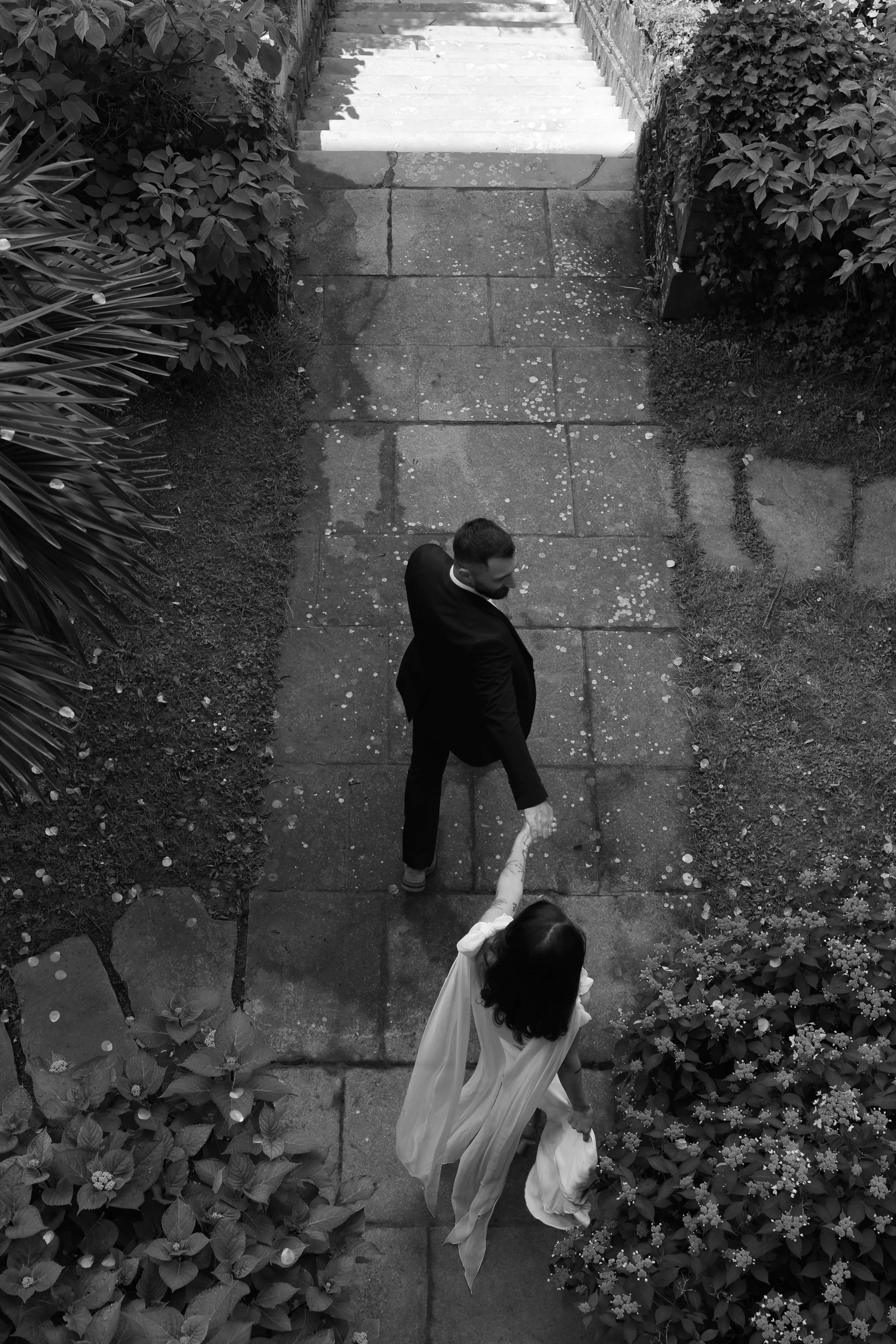 Une photo en noir et blanc prise d'en haut montrant un homme en costume marchant main dans la main avec une femme vêtue d'une robe longue, dans un jardin bordé de végétation.