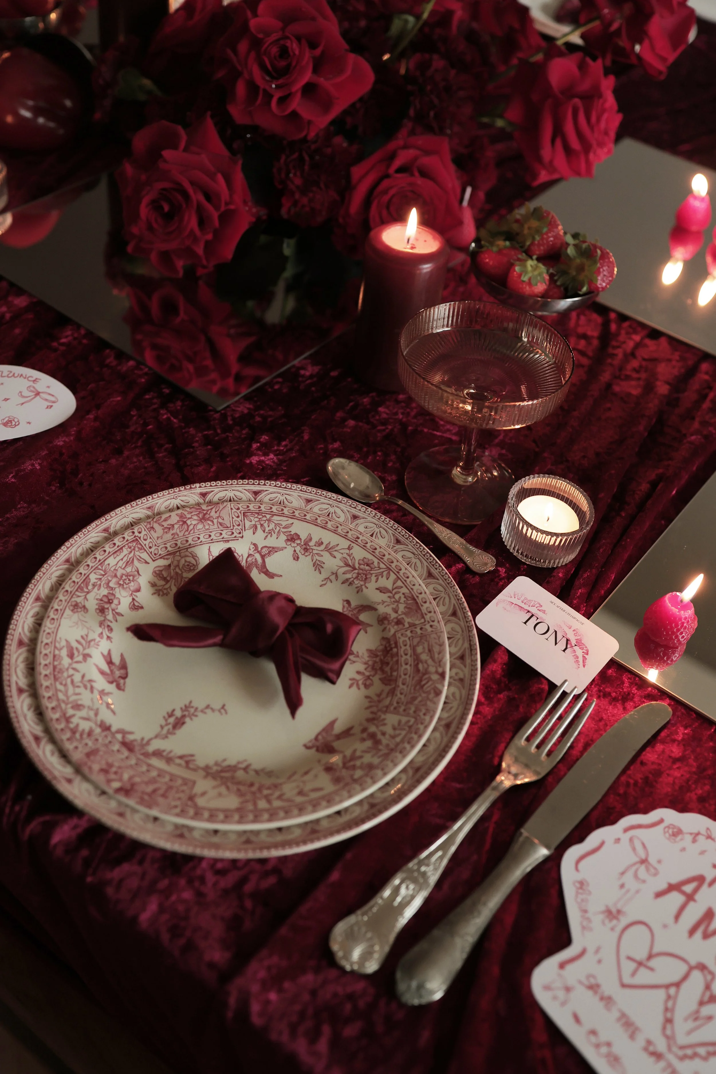 Une table élégante avec une assiette en porcelaine rose et blanche, une serviette en tissu de couleur bordeaux, des couverts en argent, une coupe à cocktail, des bougies allumées, un bouquet de roses rouges, des fraises, et une décoration raffinée da