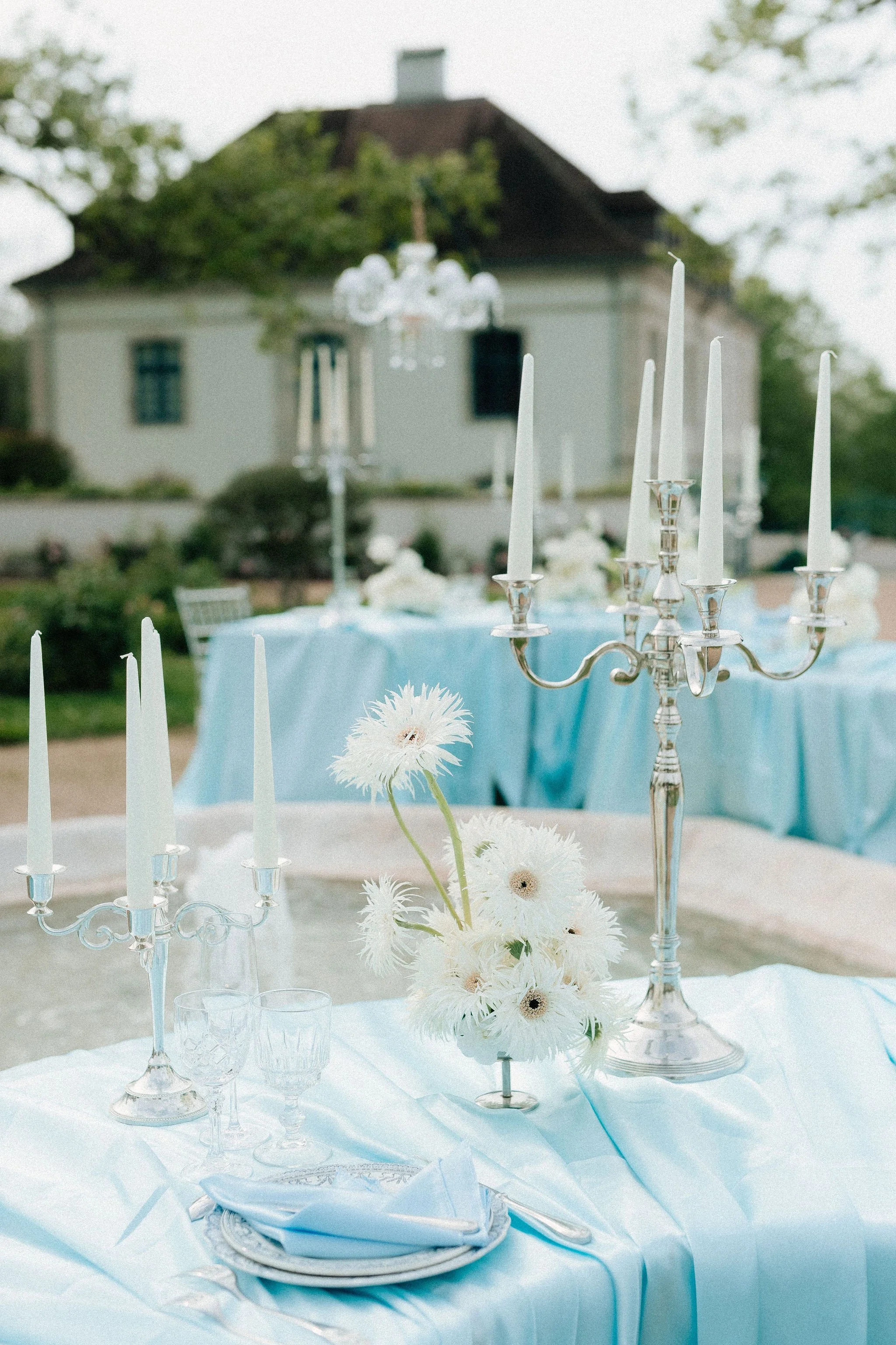 Table de fête avec une nappe bleue, des bougeoirs en argent avec des bougies non allumées, un bouquet de fleurs blanches, et une vaisselle élégante, en extérieur devant une maison avec un arbre en arrière-plan.