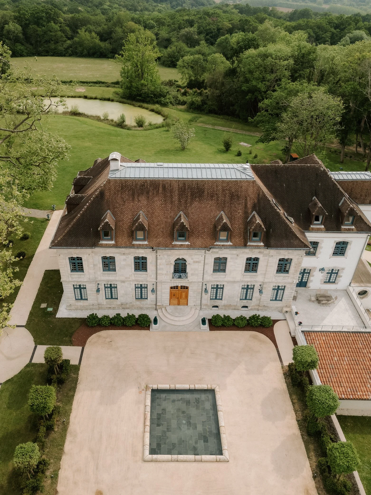 Photo aérienne d'un bâtiment ancien avec toit en tuiles rouges, fenêtres à volets bleus, devant une cour avec bassin et plantes, entouré de végétation dense et une étendue d'eau
