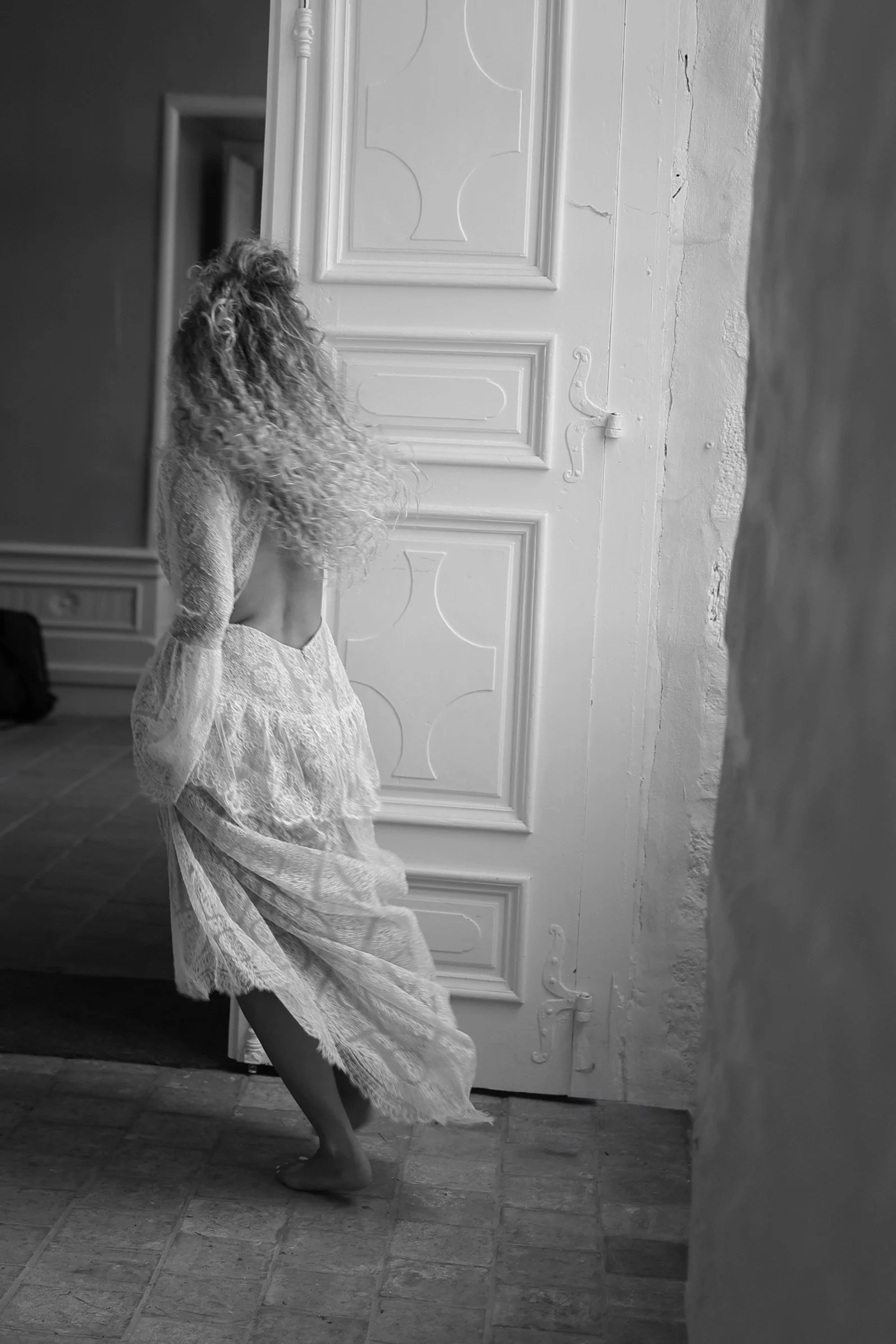 Fille en robe en dentelle blanche dans une pièce avec porte blanche et murs en pierre. La photo est en noir et blanc.