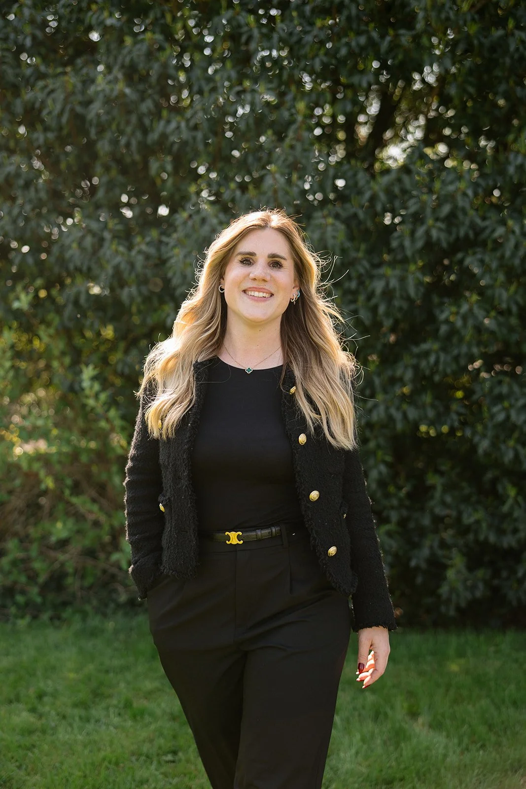 Une femme souriante avec des cheveux longs et blonds, portant un vêtement noir, posant devant un buisson vert.