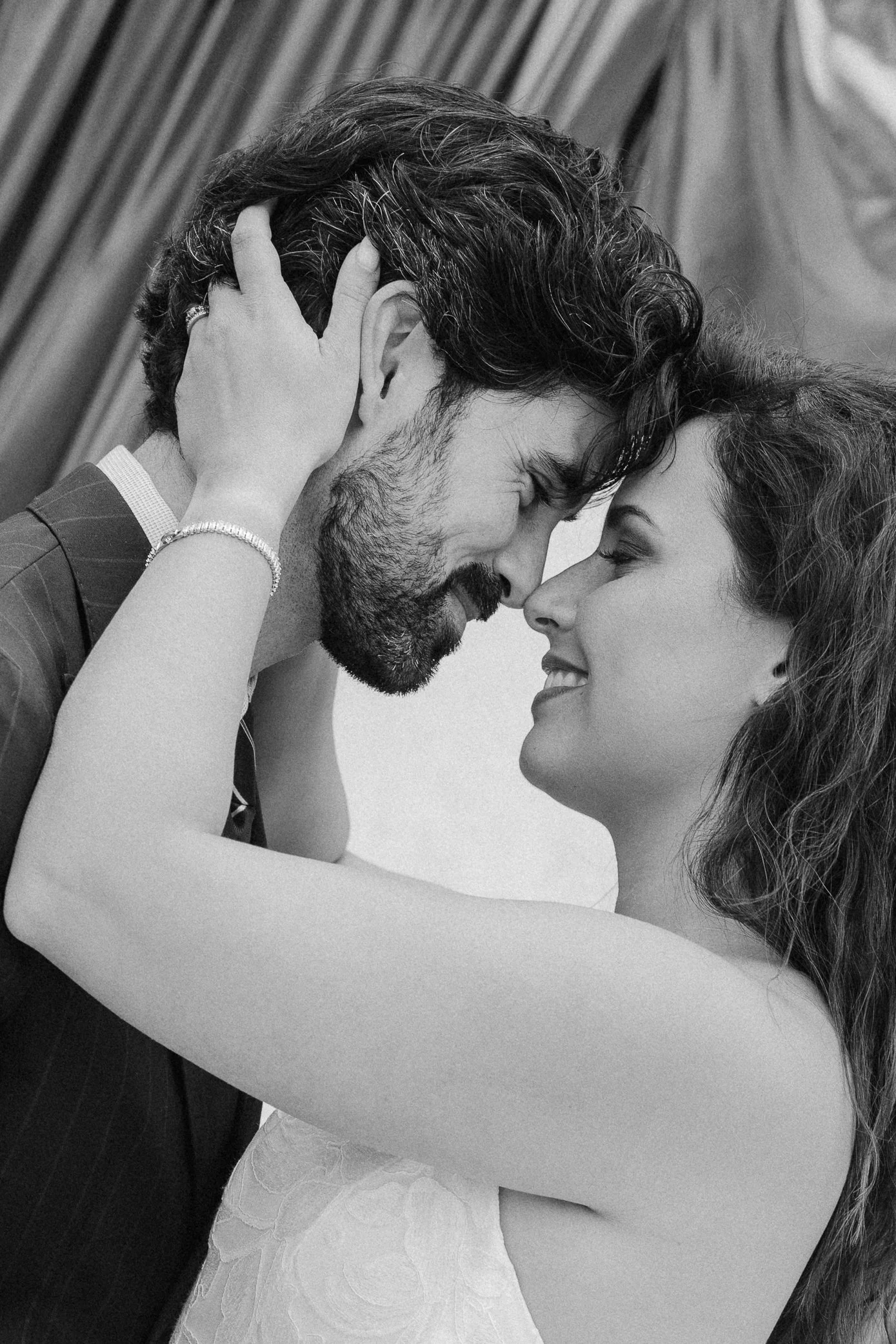 Un couple entrelace ses fronts, yeux fermés, dans une étreinte intime. La femme sourit légèrement, portant un vêtement blanc et un bracelet, tandis que l'homme a des cheveux ondulés et une barbe. Arrière-plan flou avec un rideau