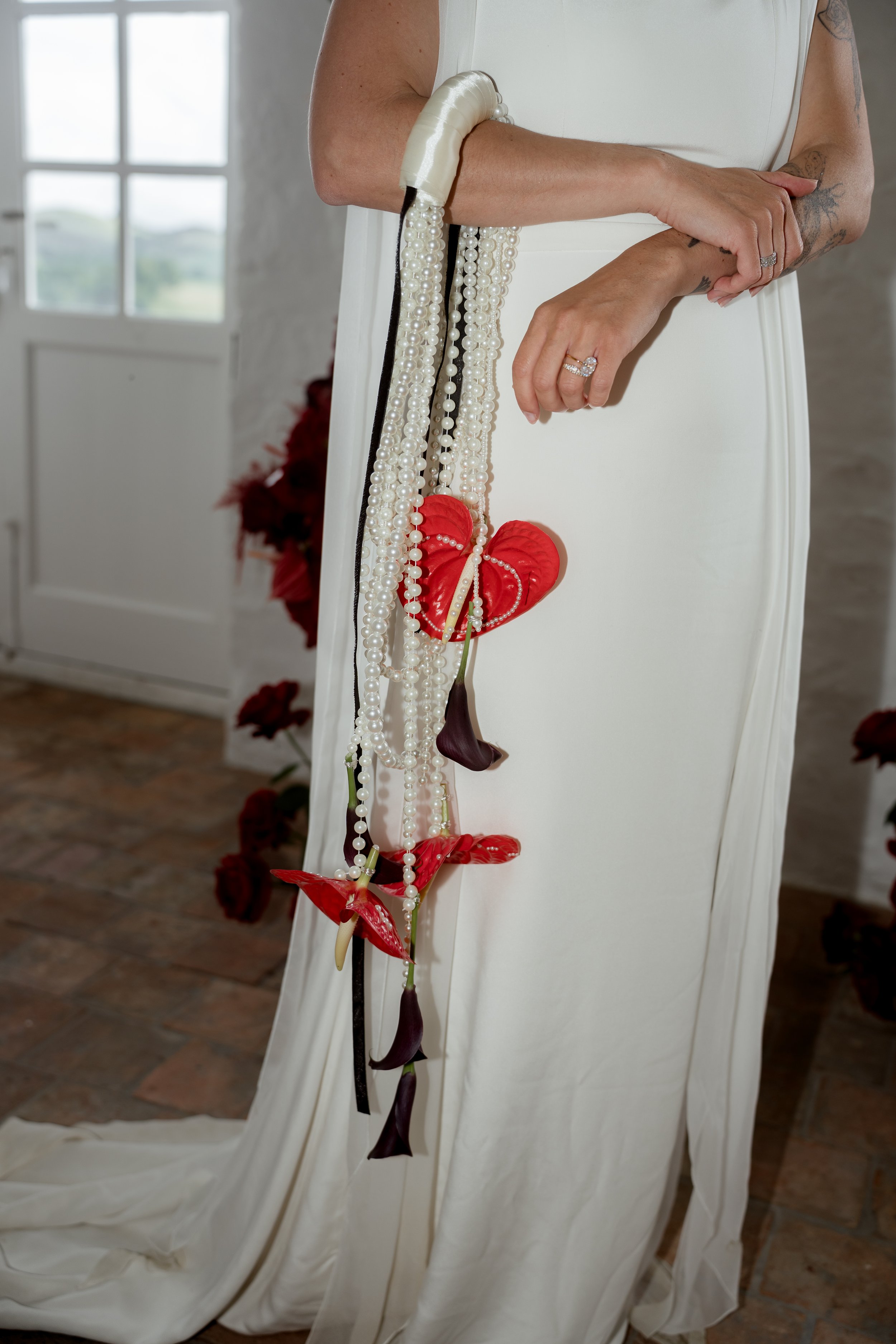 Une robe de mariage blanche, avec des bijoux de perles, et des orchidées rouges en décoration. La photo montre un détail de la robe et accessoires, avec des bras tatoués et des mains portant des bagues.