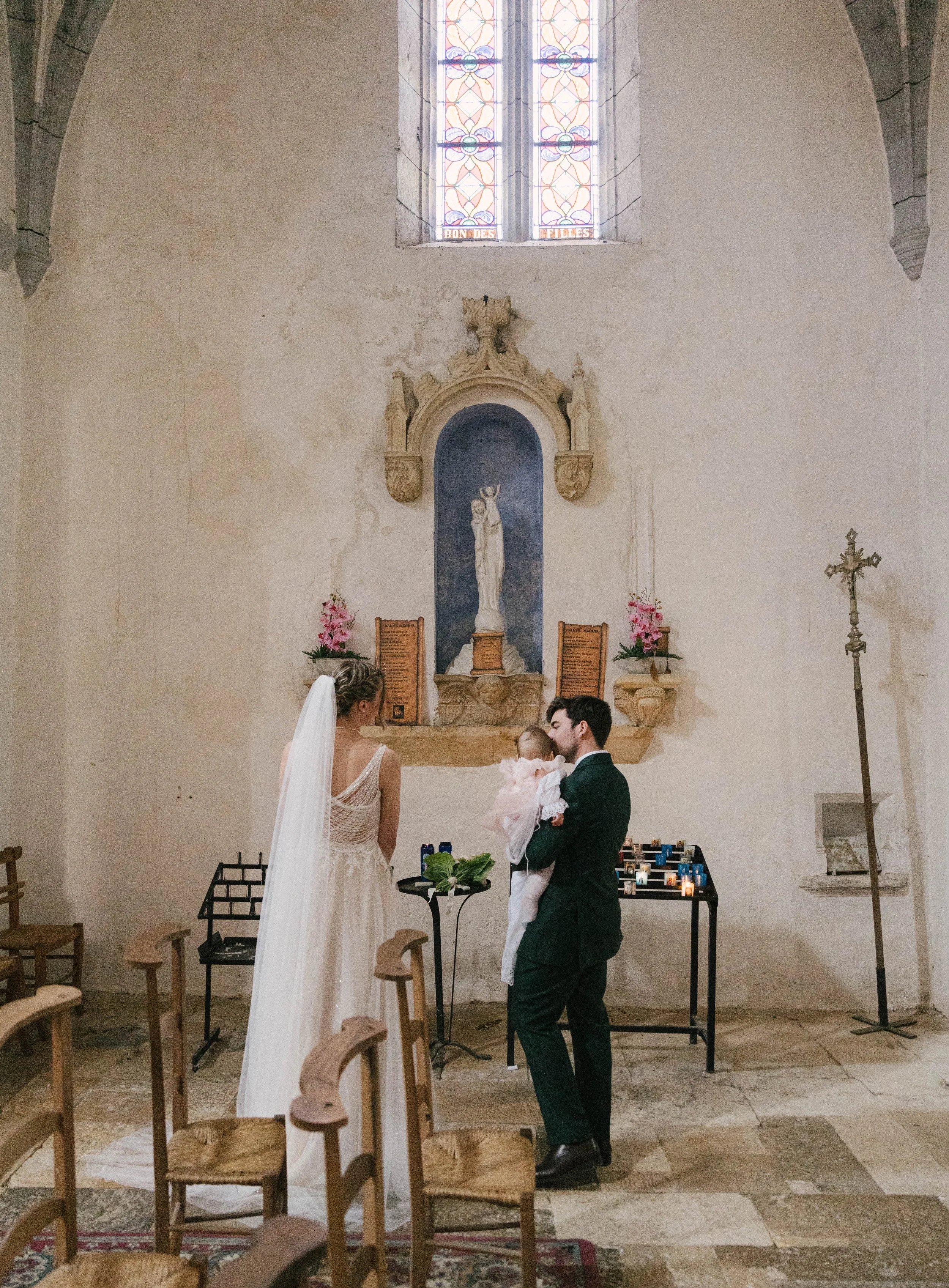 Un mariage avec un couple devant un autel dans une église. La mariée porte une robe blanche avec un voile, le marié porte un costume sombre, et ils tiennent un bébé. L'autel comporte une statue religieuse, des bouquets de fleurs et des bougies.