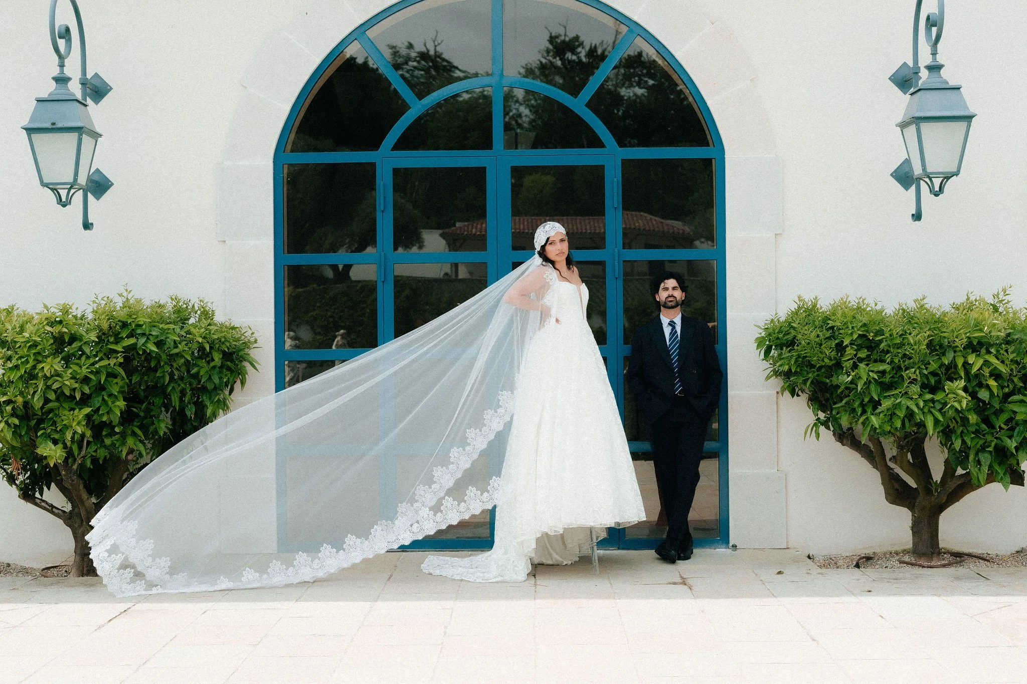 Une jeune mariée en robe blanche avec un voile long se tient devant une grande fenêtre bleue, accompagnée d'un homme en costume sombre. Il y a deux arbres taillés en forme de boule de chaque côté, et deux lampadaires suspendus, tout cela devant un mu