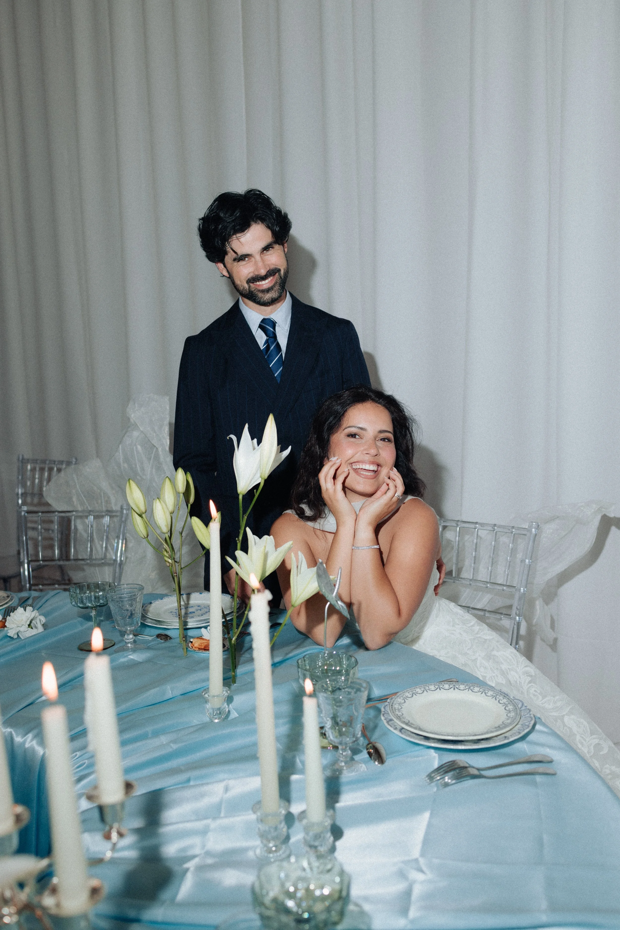 Un couple souriant à une fête, assis à une table décorée avec des fleurs blanches, des bougies et une vaisselle élégante, avec un homme en costume sombre et une femme en robe de mariée.