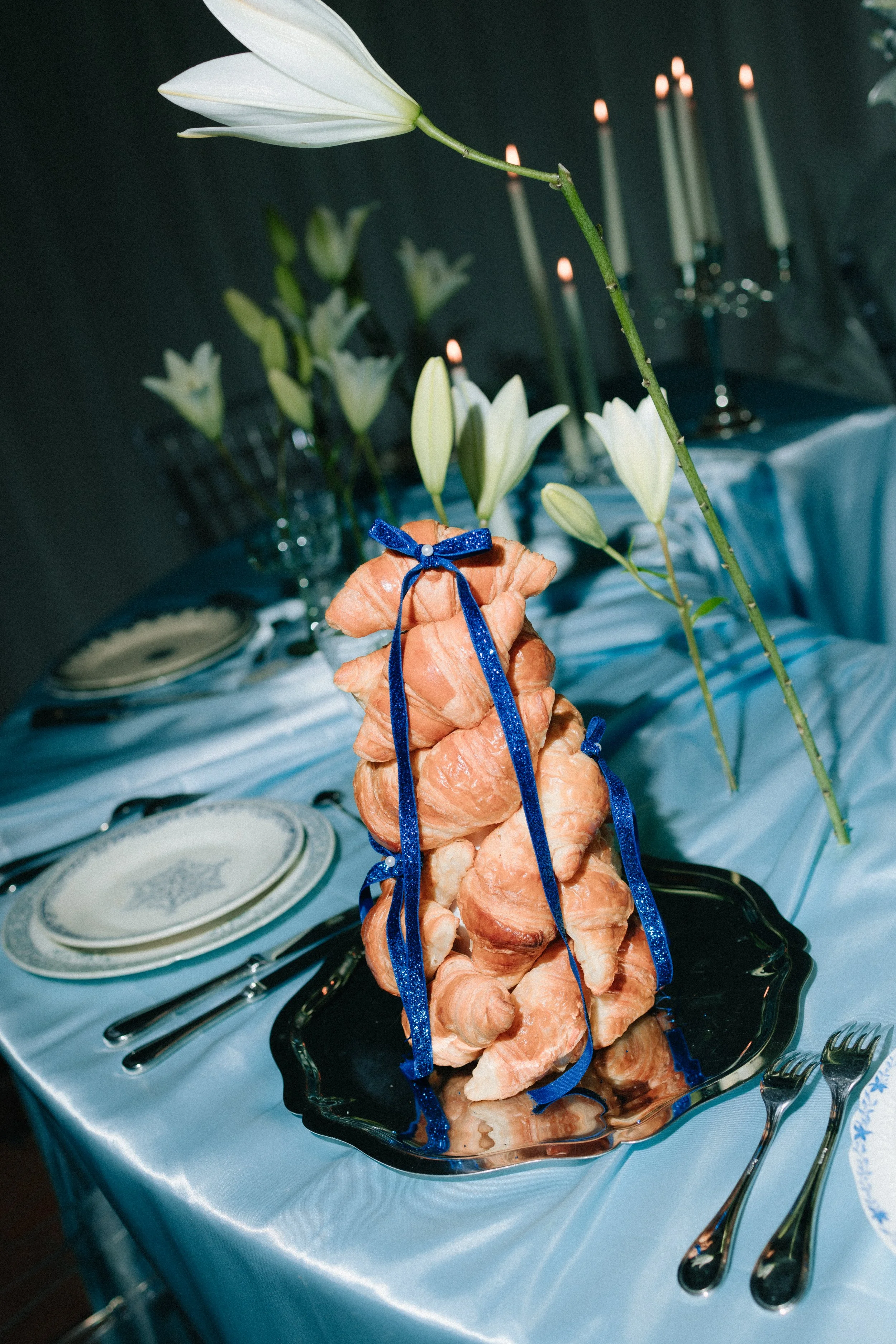 Arrivée d'un gâteau de viande en forme de torsades, décoré d'un ruban bleu, sur une table de fête avec assiettes, couverts, et fleurs blanches en fond.