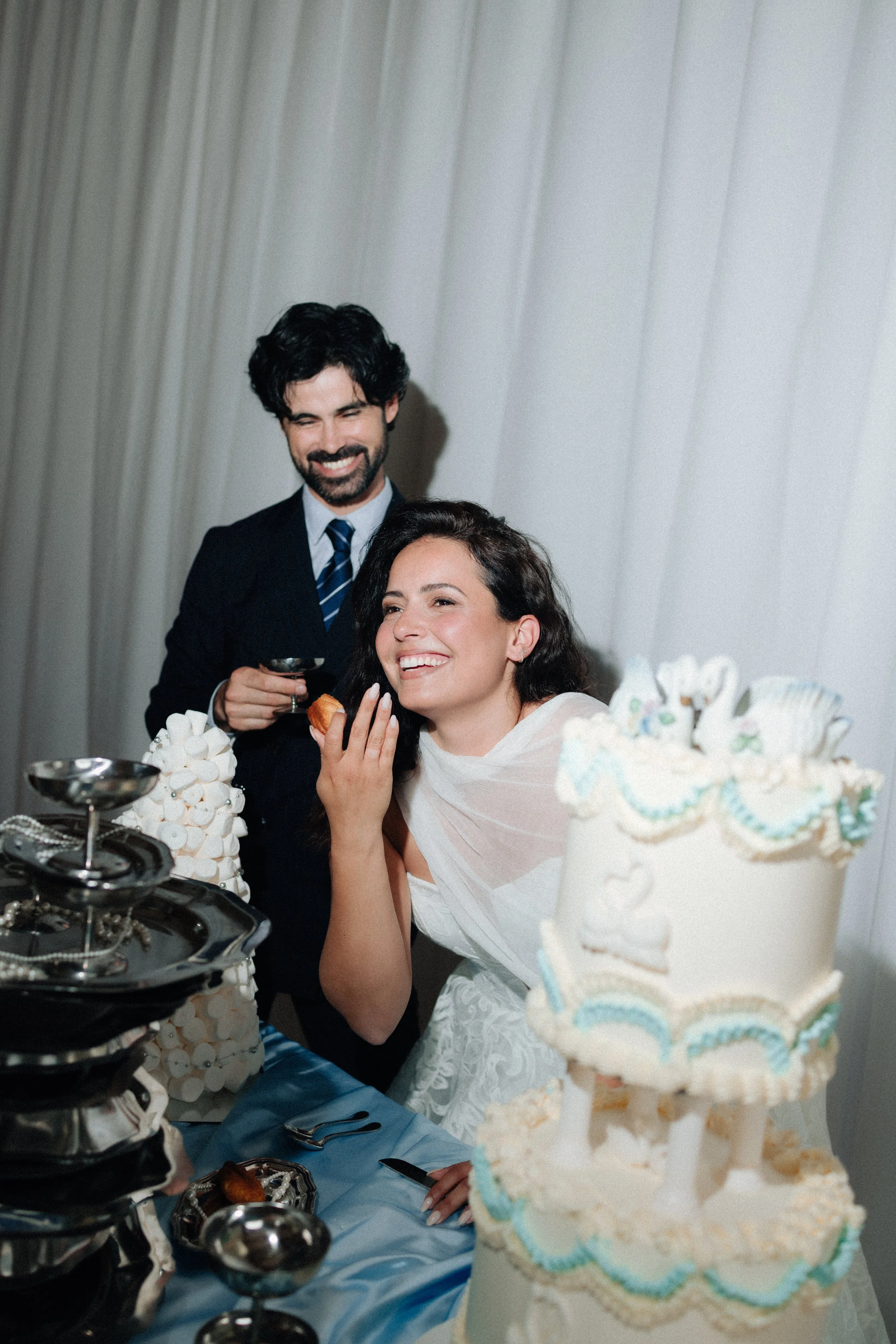 Un homme en costume souriant et une femme en robe, probablement lors d'une célébration ou d'un mariage, entourés d'un gâteau à plusieurs étages décoré de motifs colorés.