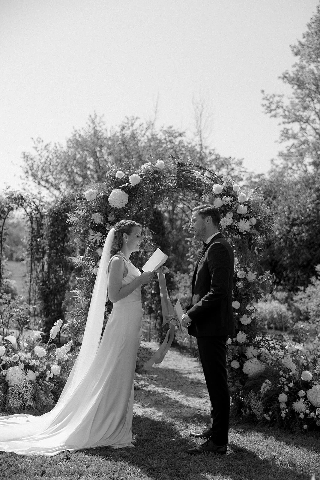 Un couple lors d'un mariage en plein air, la femme en robe de mariée lit ses vœux sous un arc fleuri, avec un décor naturel de jardin.
