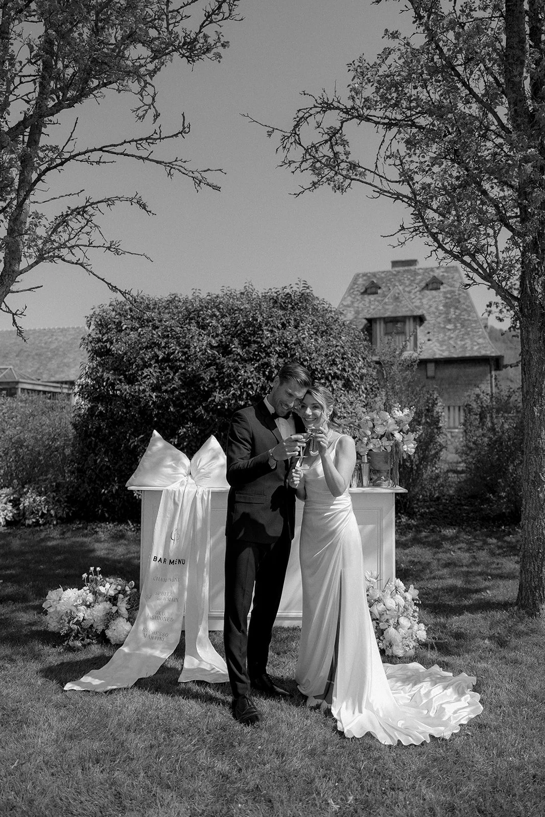 Un couple habillé en tenue de mariage se tenant côte à côte dans un jardin, levant des verres pour faire un toast, avec une table décorée de fleurs derrière eux.