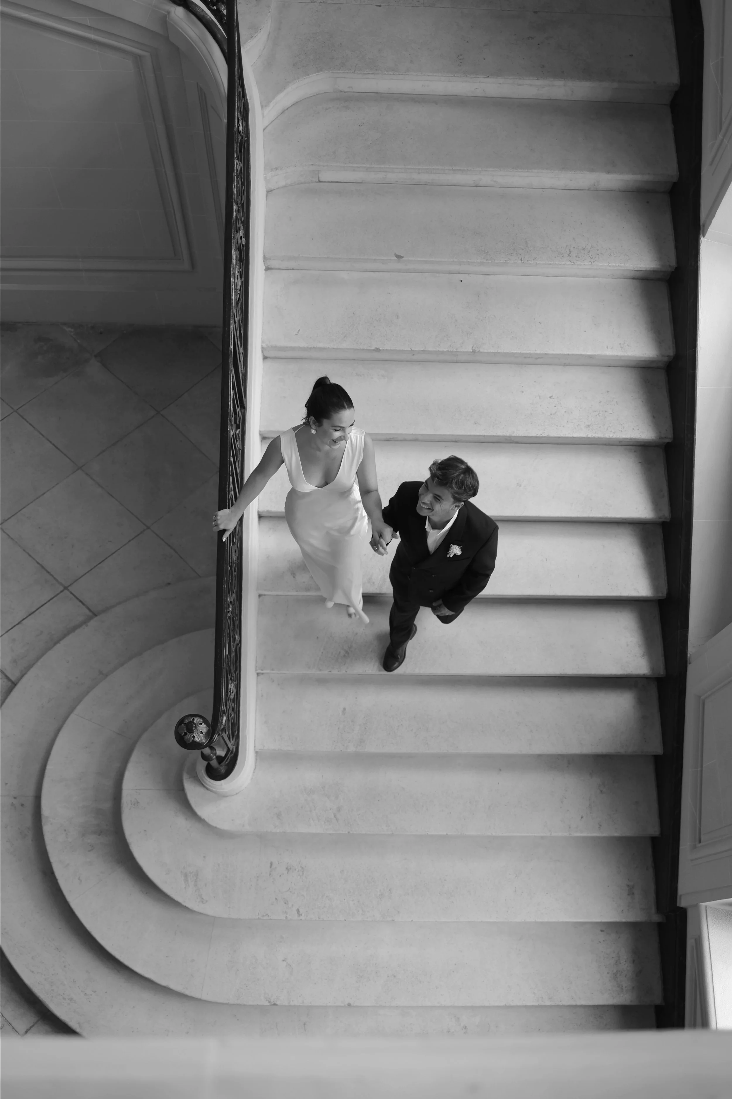 Un couple habillé en blanc et noir marchant main dans la main sur un escalier en pierre, vue prise du dessus.