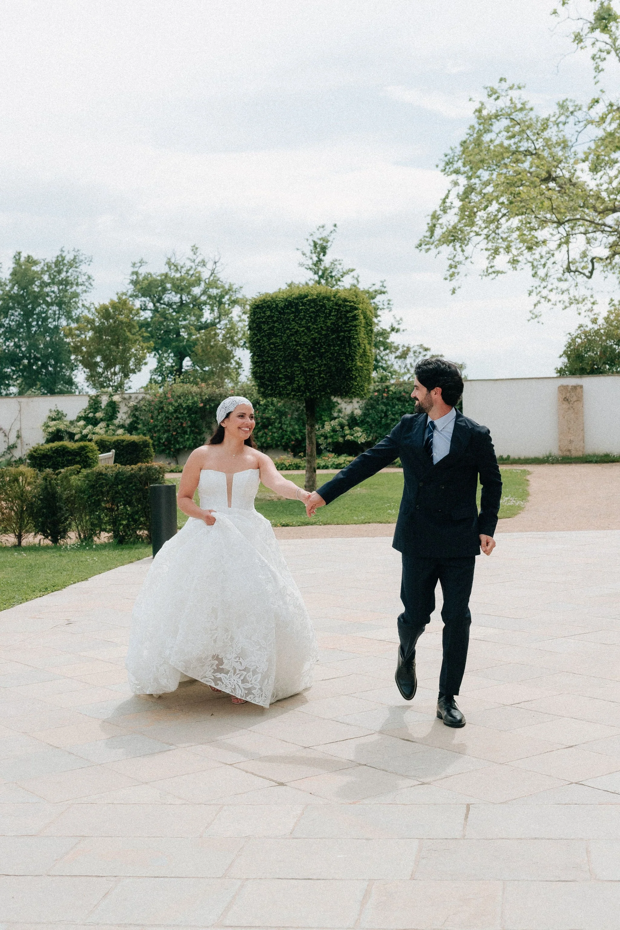 Un couple de mariés, la femme en robe de mariage blanche et le homme en costume, se tenant la main lors d'une promenade dans un jardin extérieur, souriant l'un à l'autre.