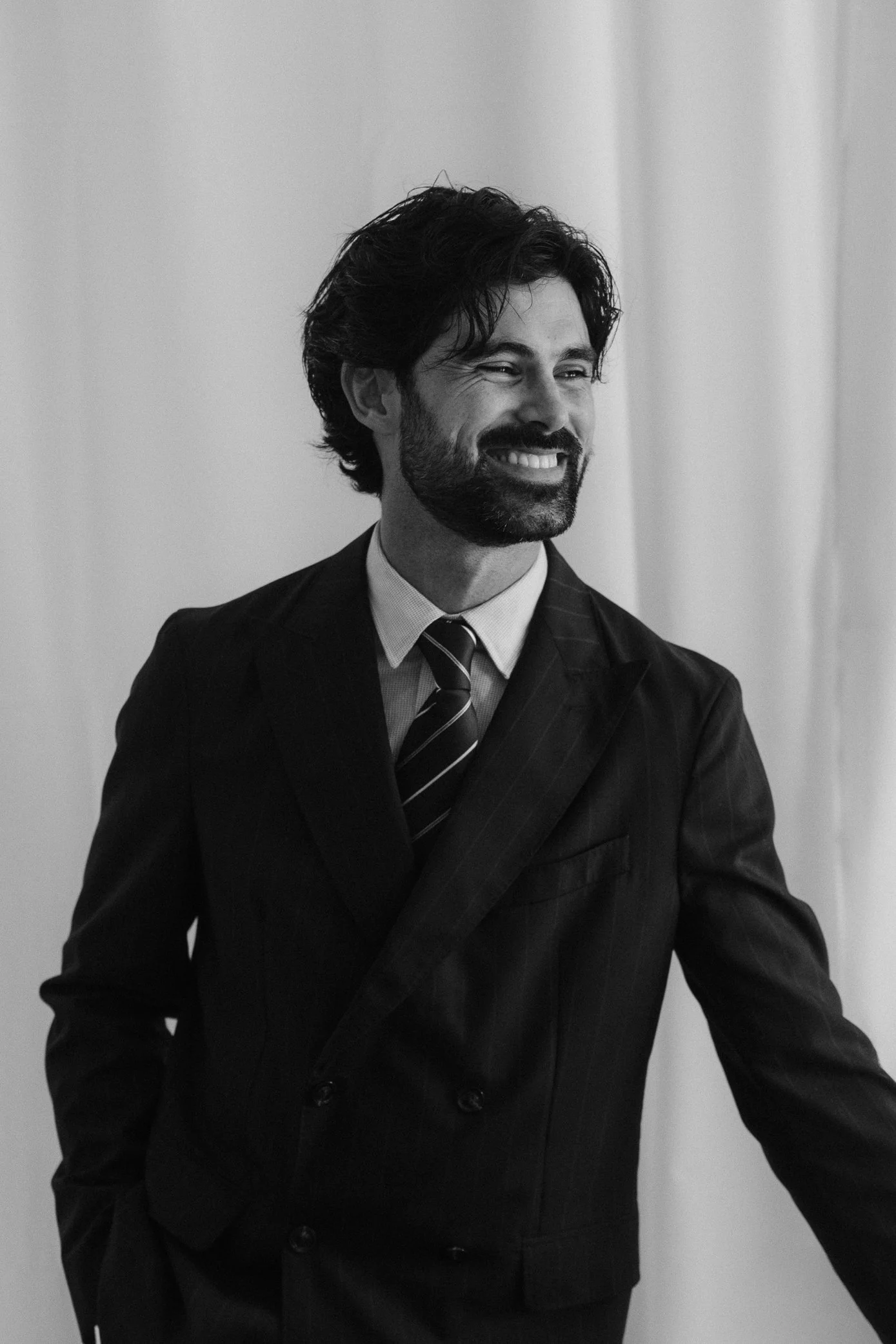 Un homme souriant, avec une barbe et des cheveux bouclés, portant un costume sombre avec une chemise claire et une cravate à rayures, dans un intérieur.