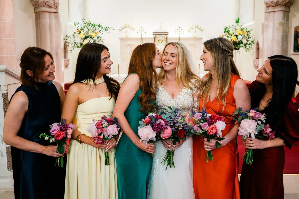 Bridemaids in multi-coloured dresses kissing bride in church