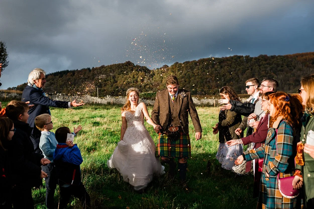 Bride and groom walking through confetti on hill overlooking Byre at Inchyra