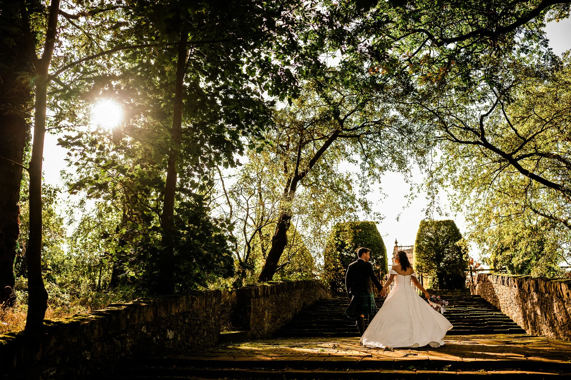 Aimee and Matthew walk along a sunlit tree-lined path at Kinmount House.