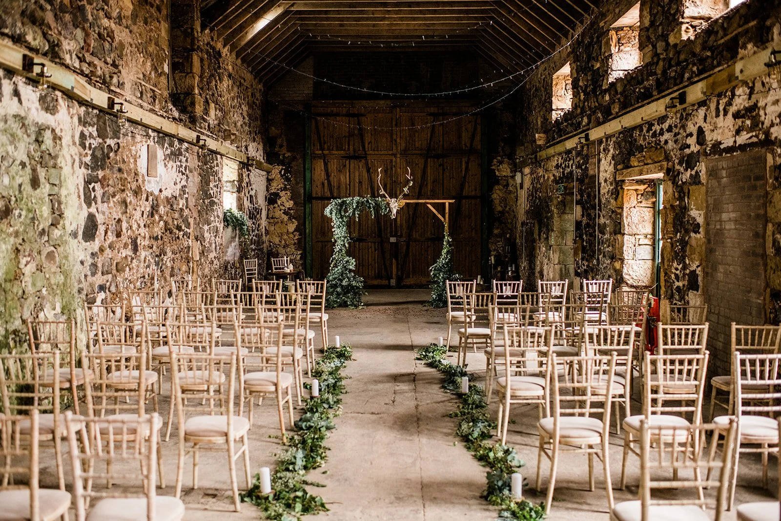 Chairs and greenery line the aisle inside the stone-walled wedding ceremony barn.